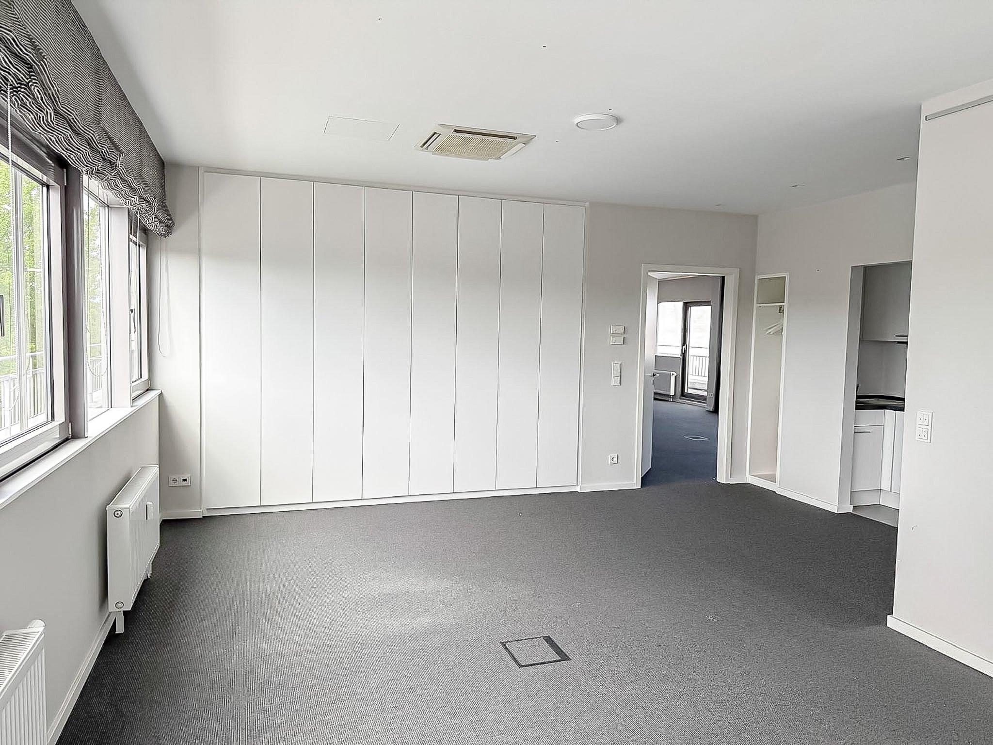 Ein leeres modern eingerichtetes Zimmer mit weißen Wänden, großen Fenstern mit schwarzen und weißen Vorhängen, grauem Teppichboden, einer weißen Heizung und einer Deckenklimaanlage.
