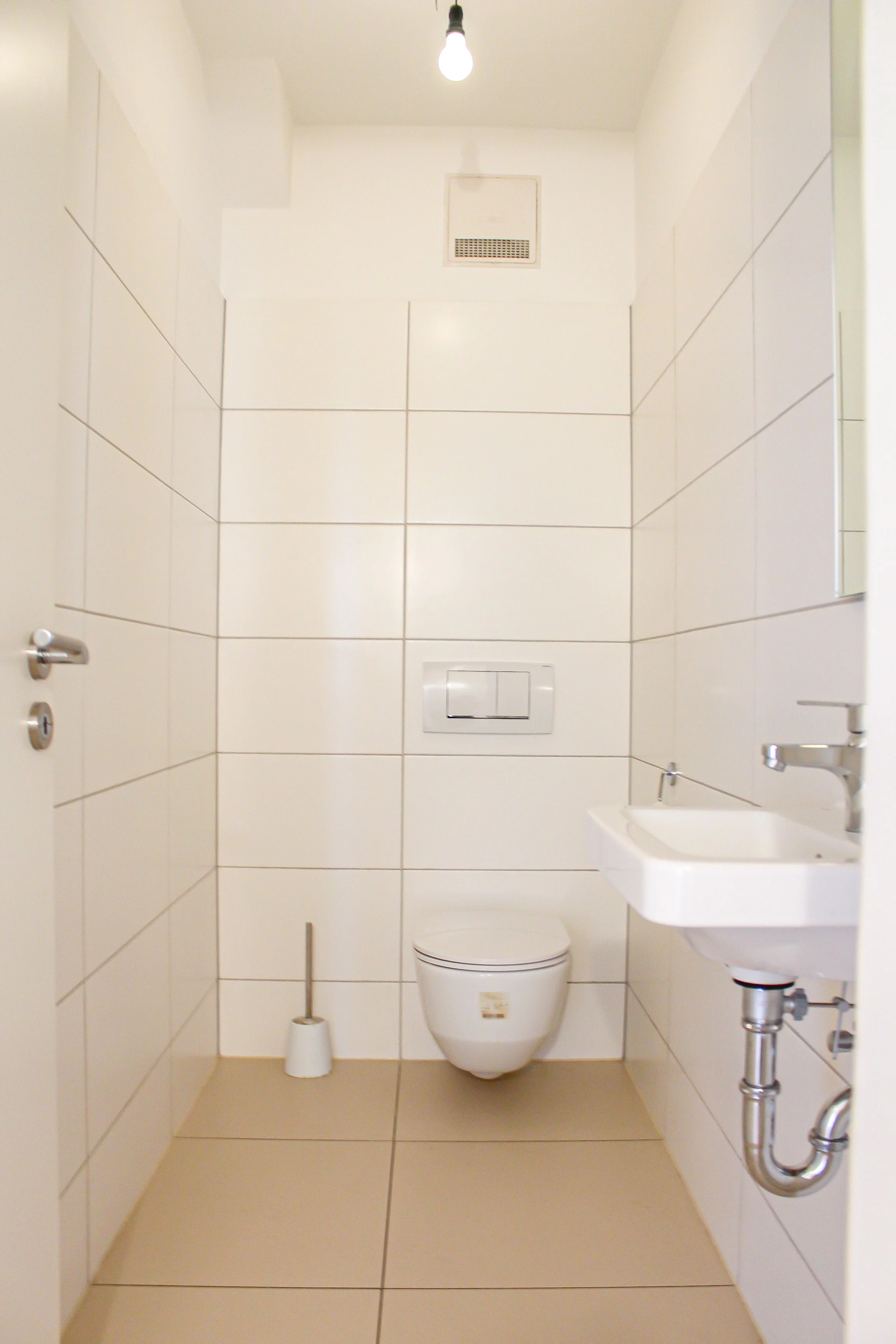 Ein kleines, weißes Badezimmer mit braunen Bodenfliesen, einer Toilette, einem kleinen Waschbecken, einem Abwischbesen und einem Pinkelhaken. Die Wände sind mit großen weißen Fliesen bedeckt, es gibt eine Deckenleuchte und einen kleinen Lüftungsschac