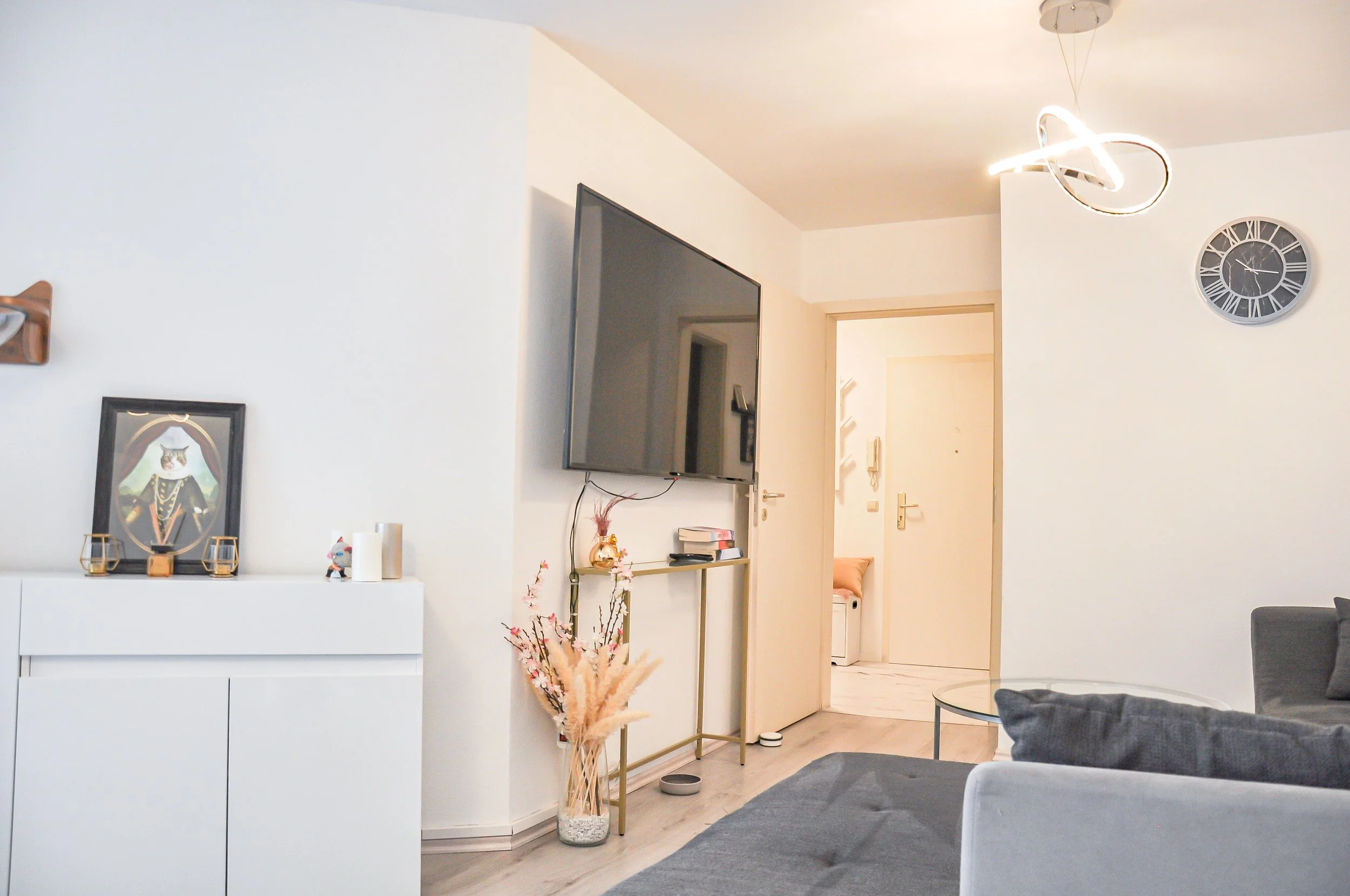 Innenansicht eines modernen Wohnzimmers mit weißer Wand, Fernseher, Wanduhr, Dekorationen und einem grauen Sofa.