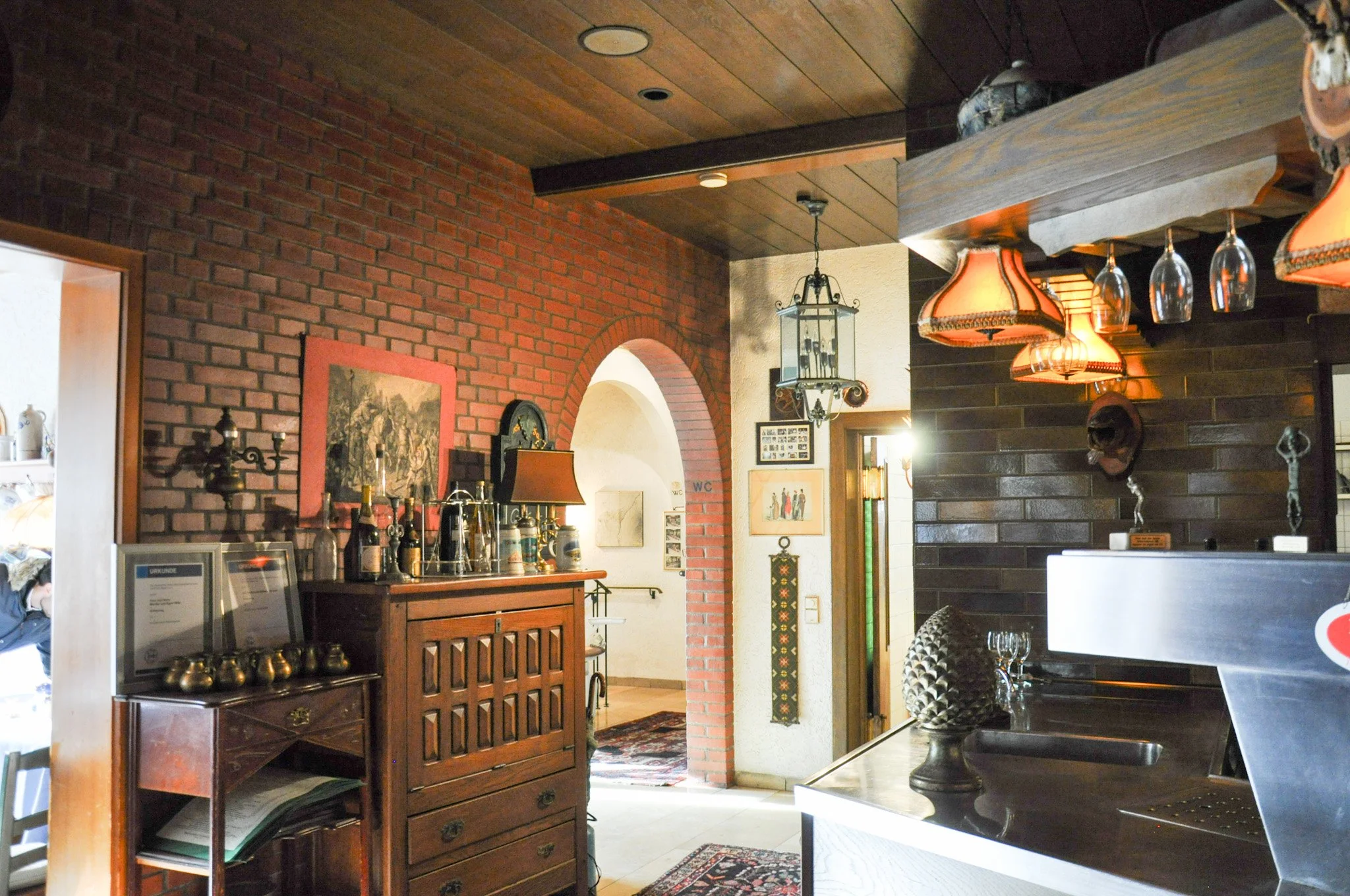 Ein Innenraum mit roten Backsteinwänden, dunklem Holzdecke, dekorativen Lampen, Kunstwerken an den Wänden und Holzmöbeln, wahrscheinlich eine Bar oder ein Restaurant.