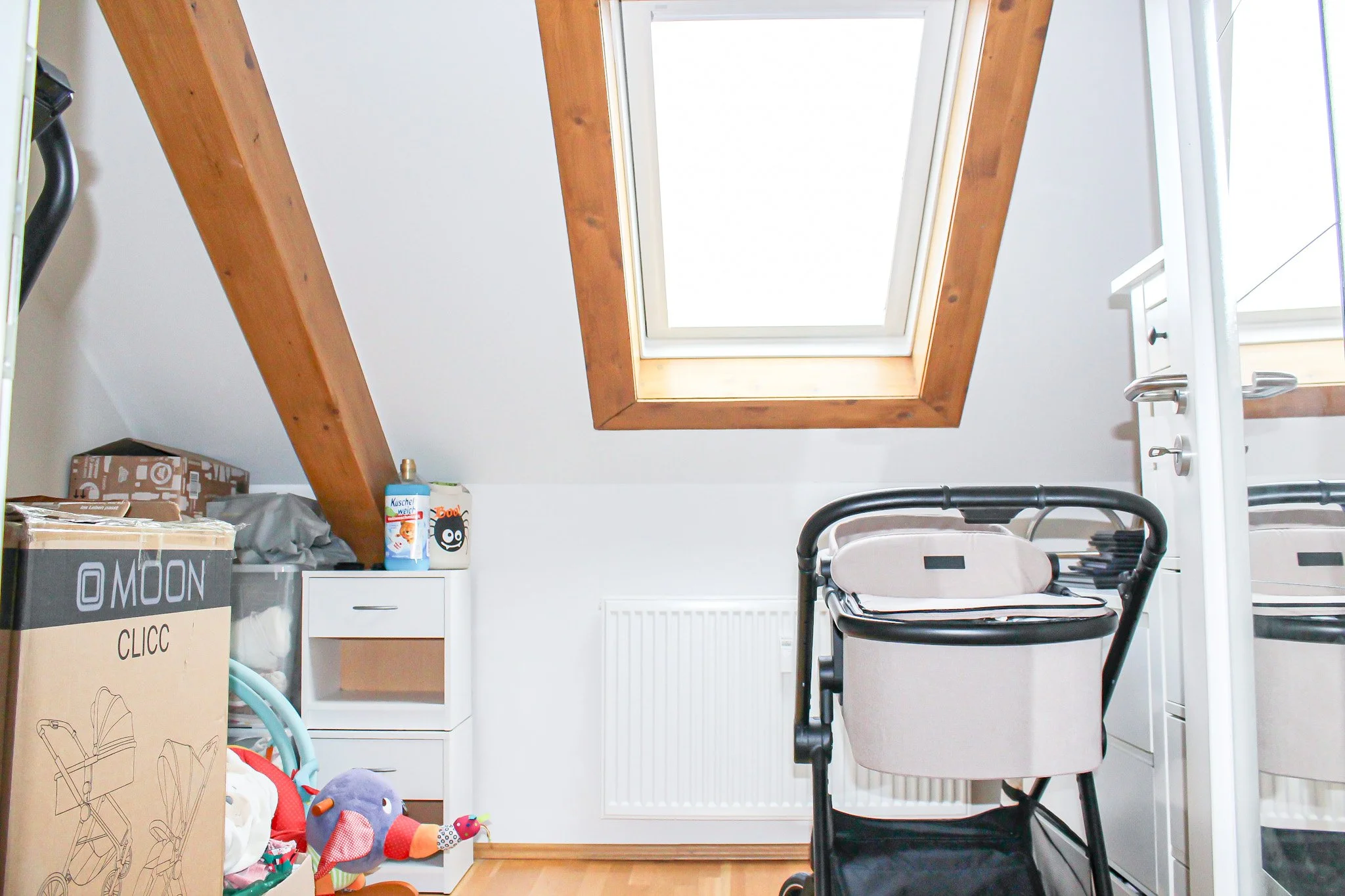 Ein Dachboden mit einem großen Dachfenster, einigen Umzugskartons, Spielzeug und Haushaltsgegenständen, die auf einem Regal und einer Kommode stehen, sowie einer weißen Heizung unter dem Fenster.