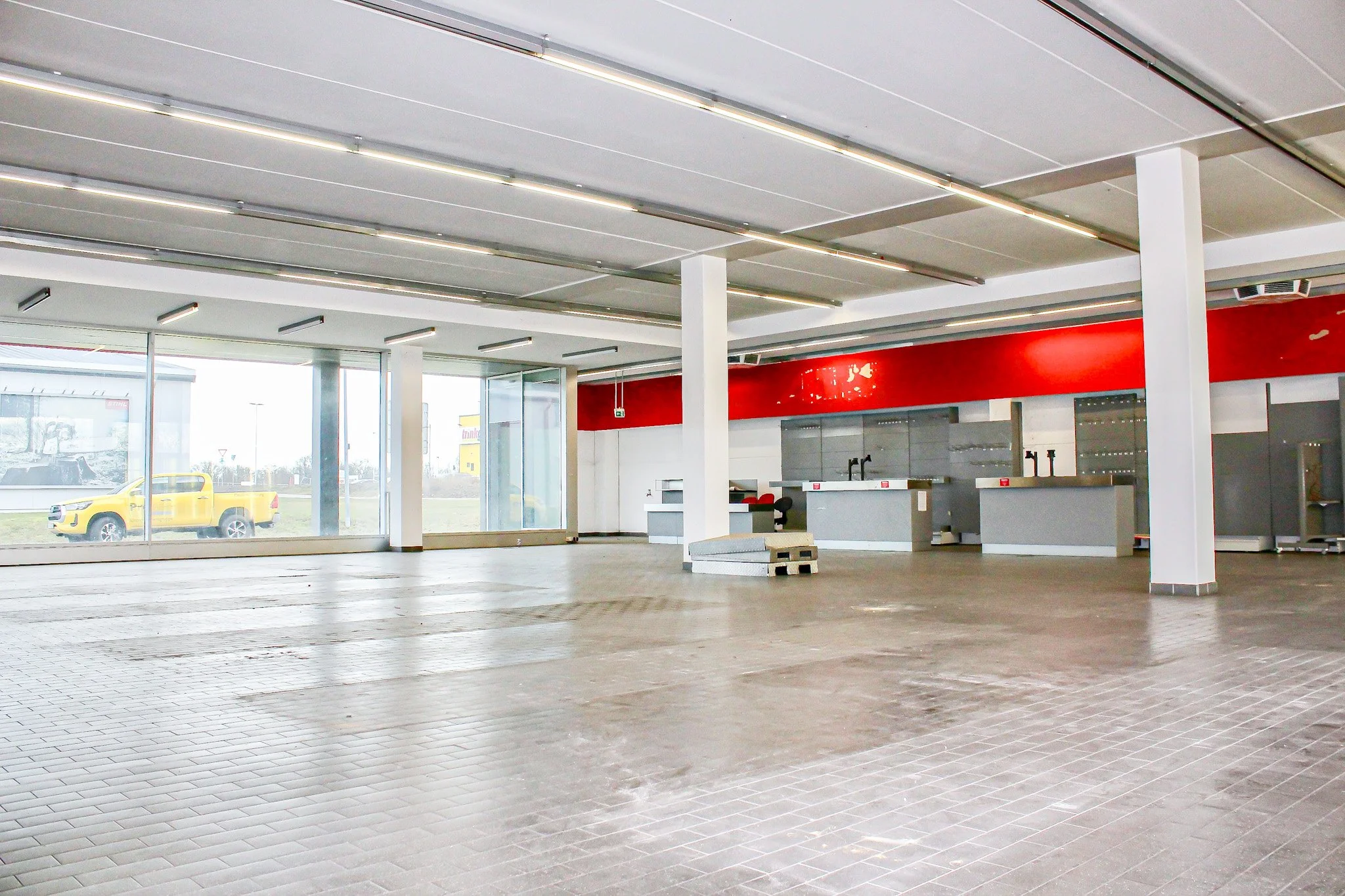 Innenansicht eines leeren Showrooms mit großen Glasfenstern, Fliesenboden, weißen Wänden, roten Akzenten an der Wand und einigen Ausstellungsregalen im Hintergrund.