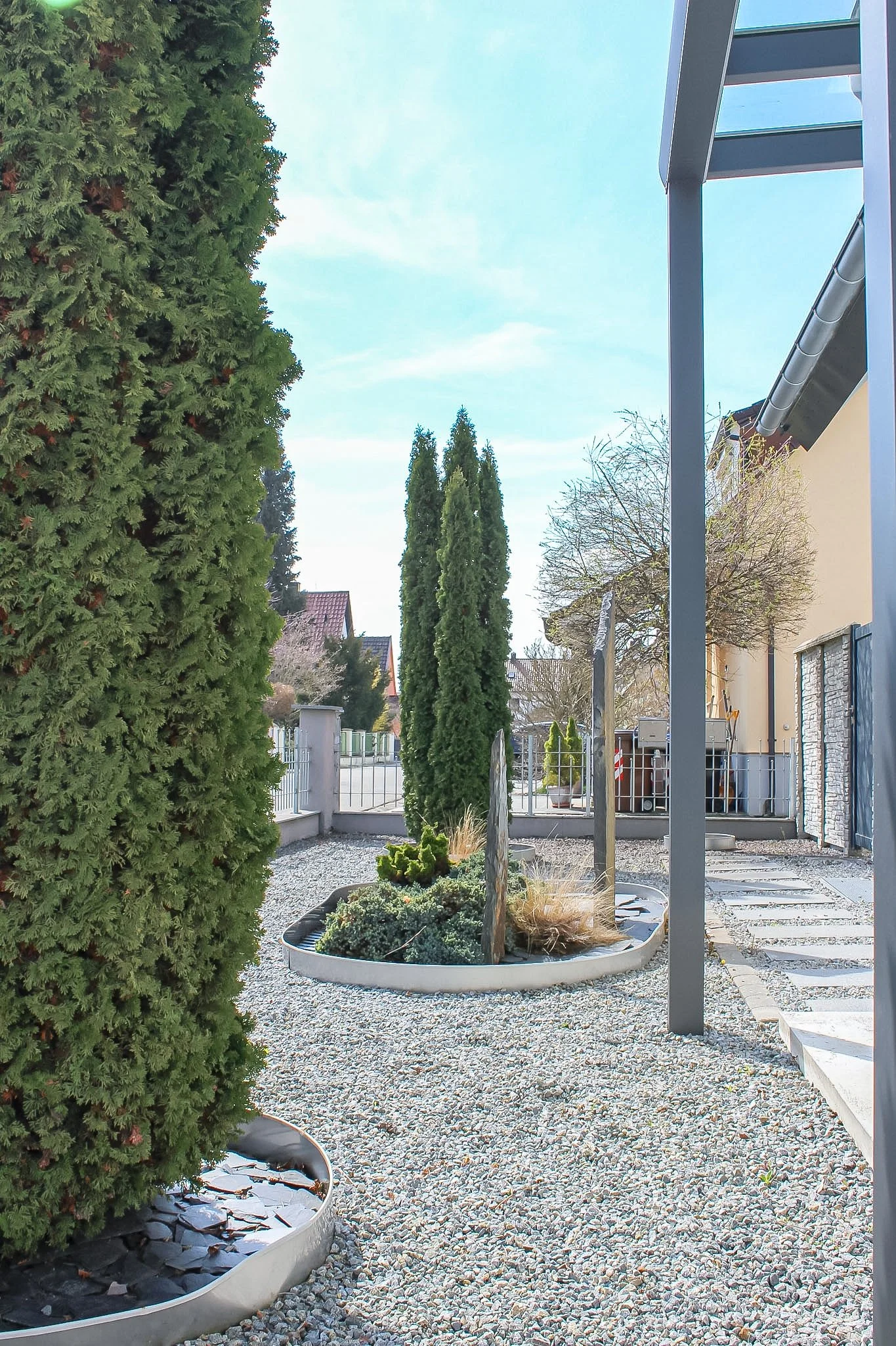 Ein gepflegter Garten mit einer großen Hecke auf der linken Seite, mehreren hohen Bäumen in der Mitte und einem modernen Haus im Hintergrund. Der Boden ist mit kleinen weißen Kieseln bedeckt, und es gibt mehrere runde Beete mit Pflanzen. Ein kleines 