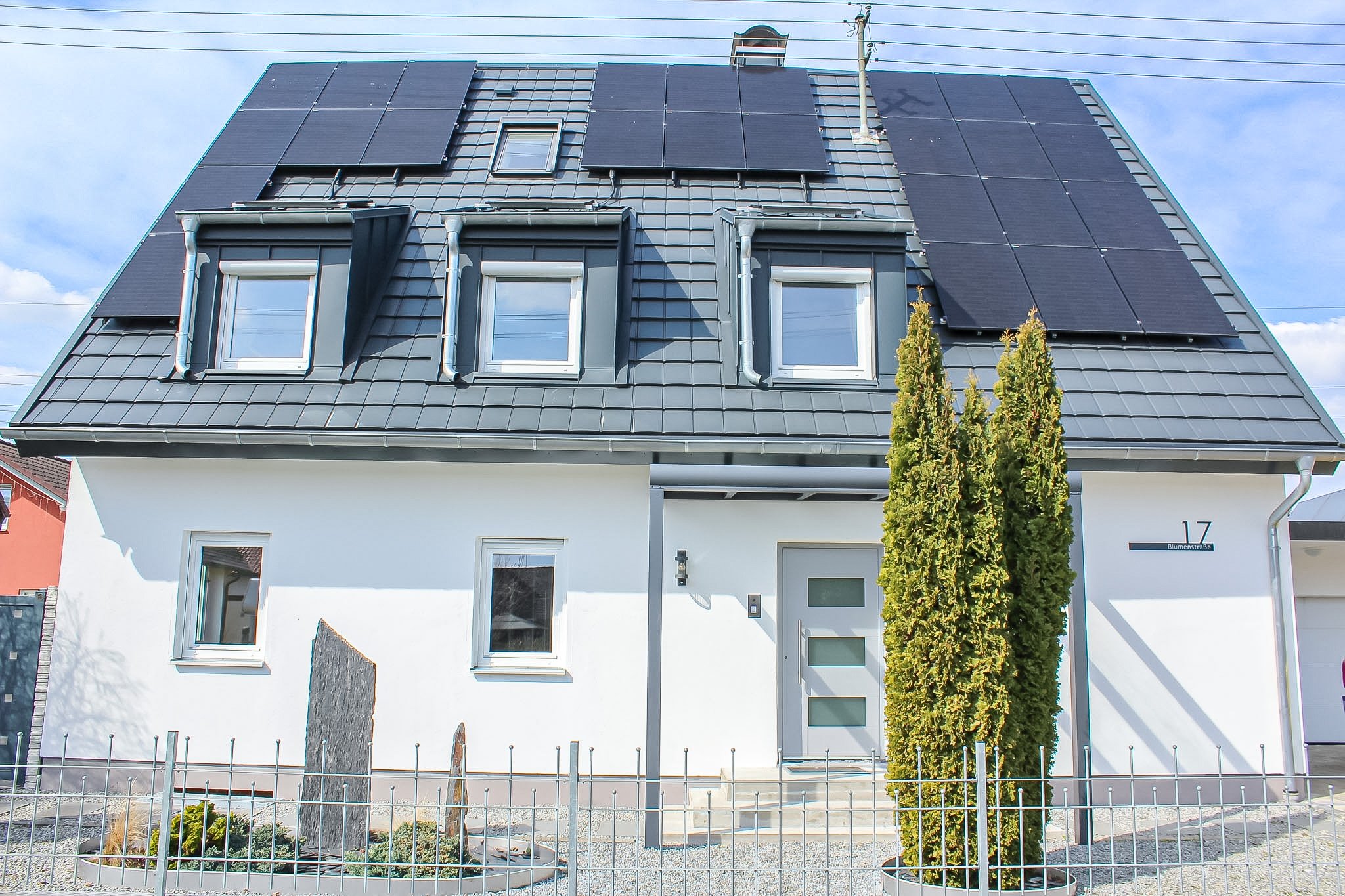 Haus mit PV-Anlage auf dem Dach, drei Dachfenster, Baum in Vordergrund, Zaun vor dem Haus, Straßenschilder.