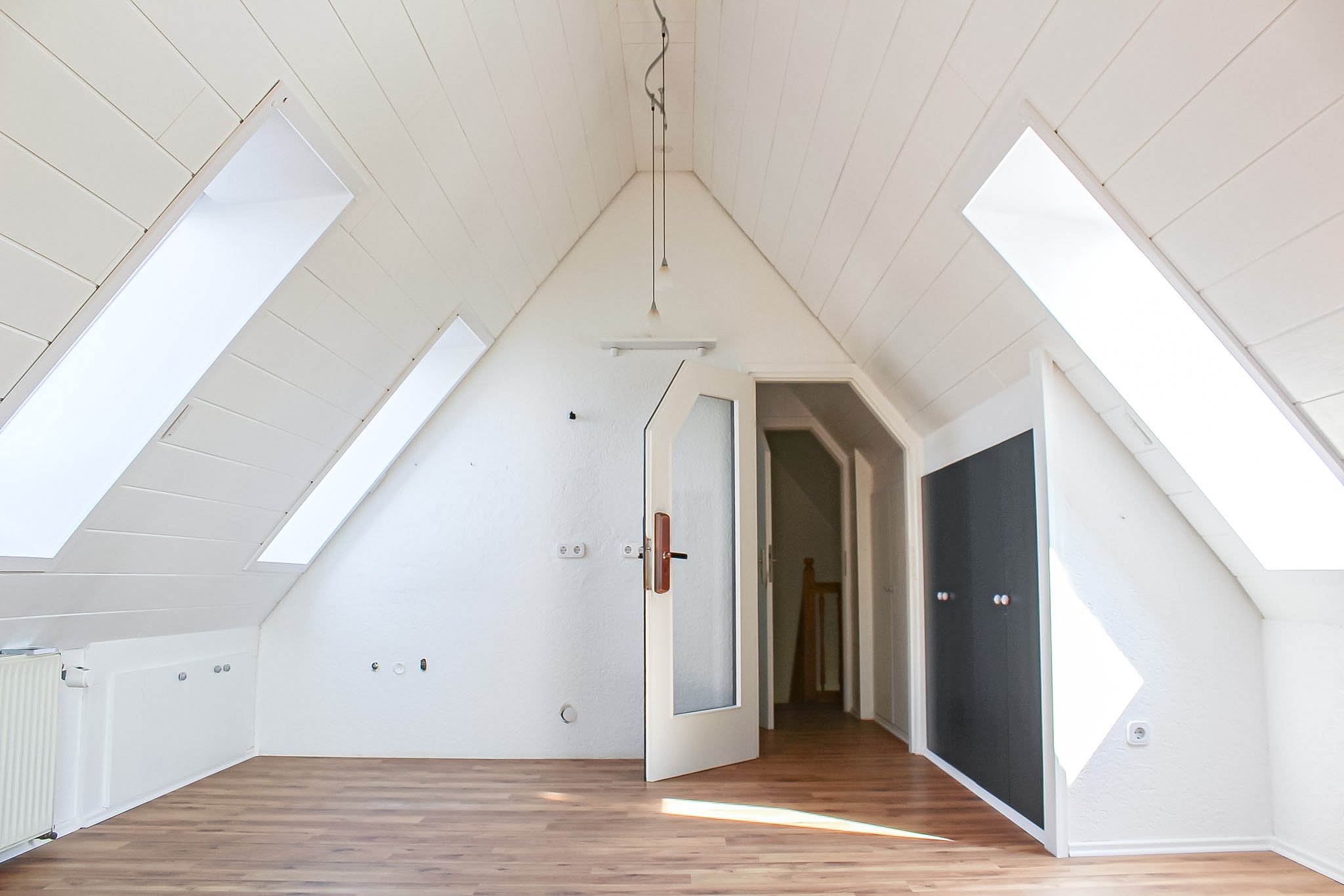 Ein Dachboden mit weißen Wänden und Deckenpaneelen, drei große Dachfenster, ein offener Türrahmen und Holzboden, viel natürliches Licht.