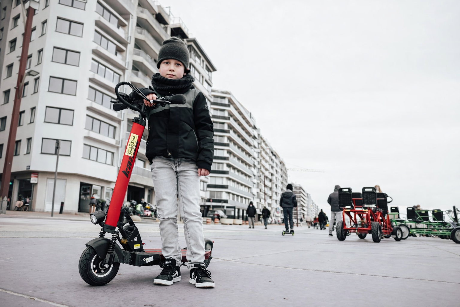 Jongetje met zwarte jas, grijze broek en pet op elektrische step in een stadsplein met meerdere mensen en moderne appartementen.