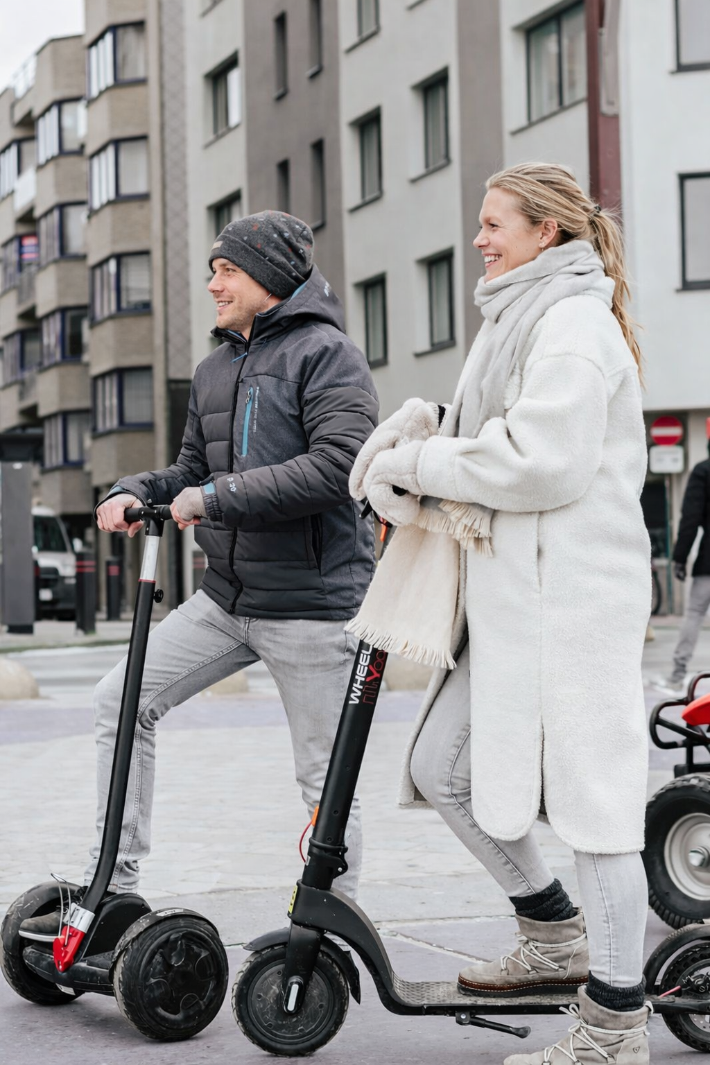 Man en vrouw rijden samen op elektrische steps in een stedelijke omgeving, beiden lachen en dragen warme kleding.