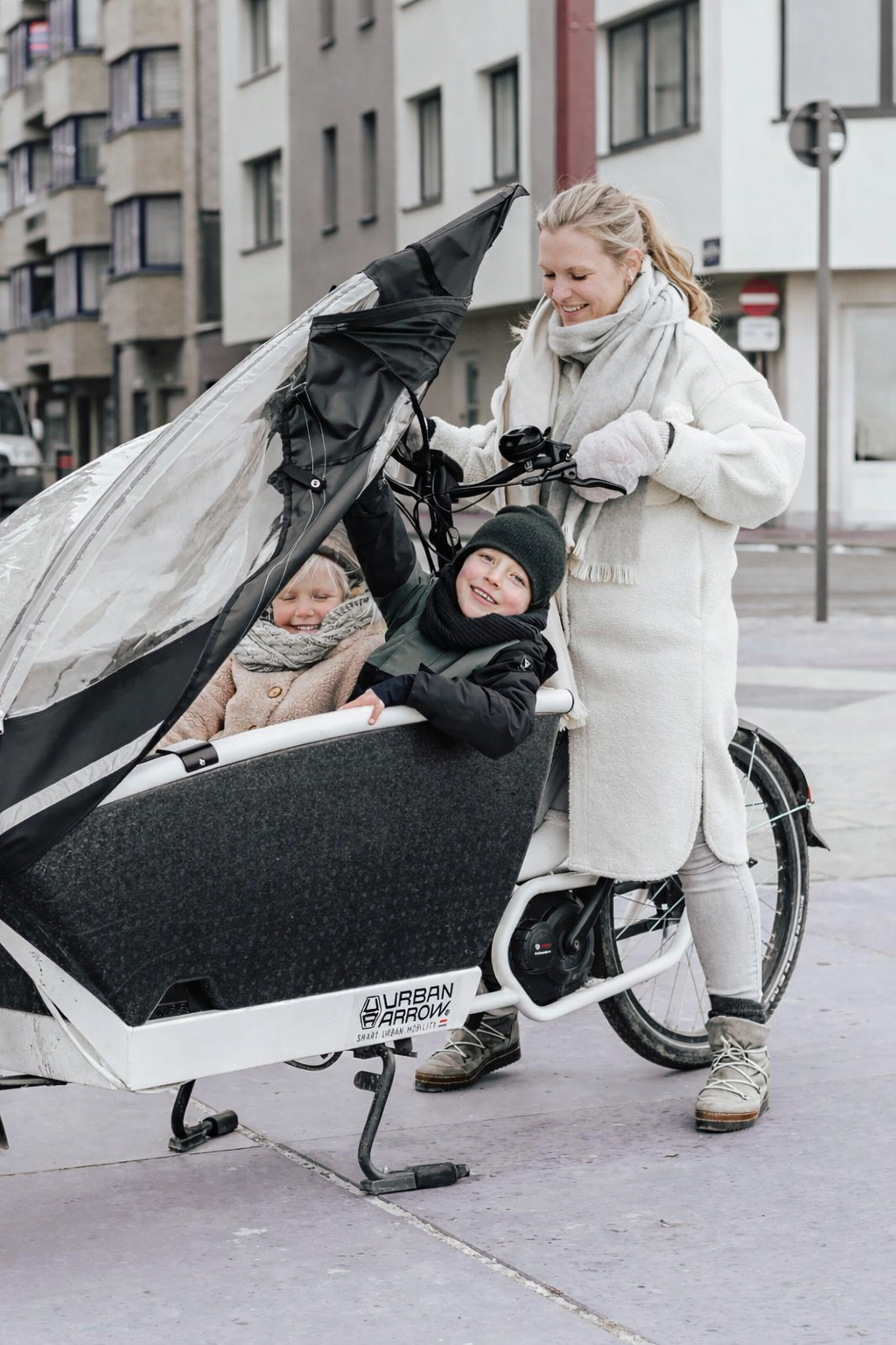 Vrouw met bakfiets, waarin twee lachende kinderen zitten, op een stedelijk plein.