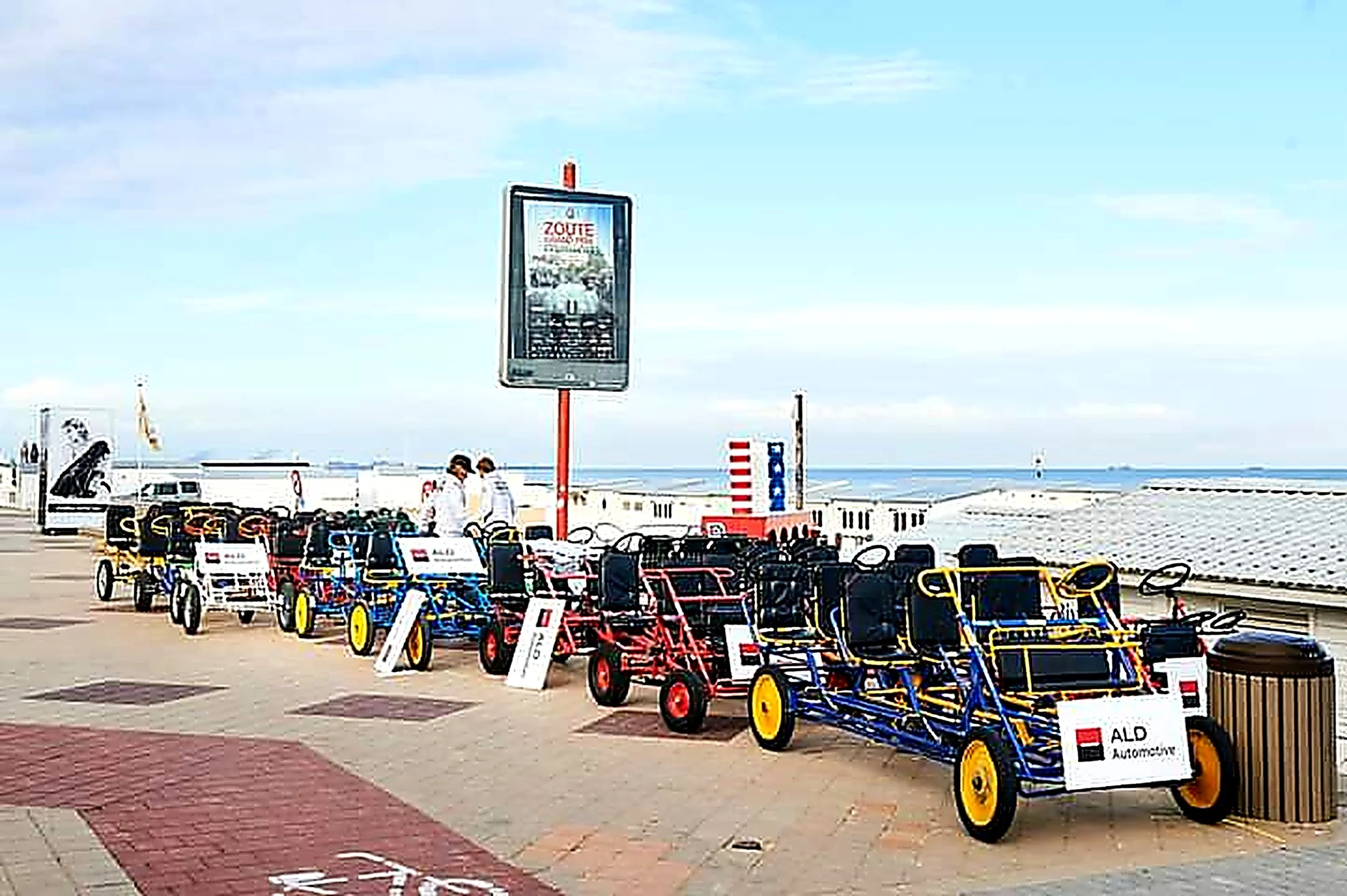 Verschillende kleurrijke gocarts geparkeerd langs de promenade met een digitaal reclamebord en de zee op de achtergrond.