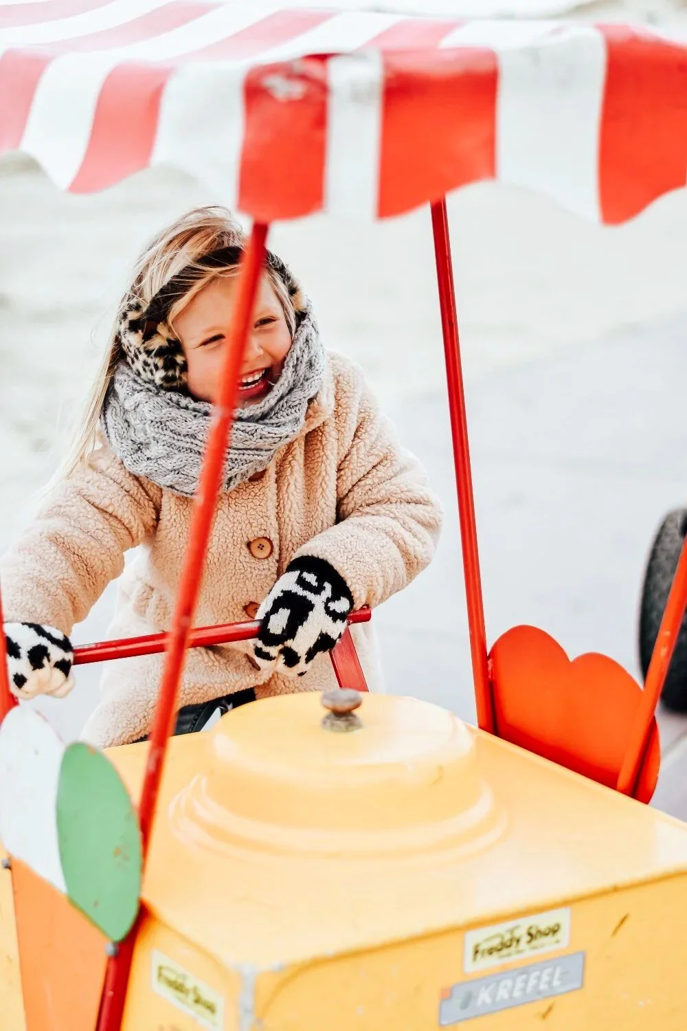 Een blij meisje in een beige jas en dierenprint handschoenen, spelen met een kindergocart op het strand.