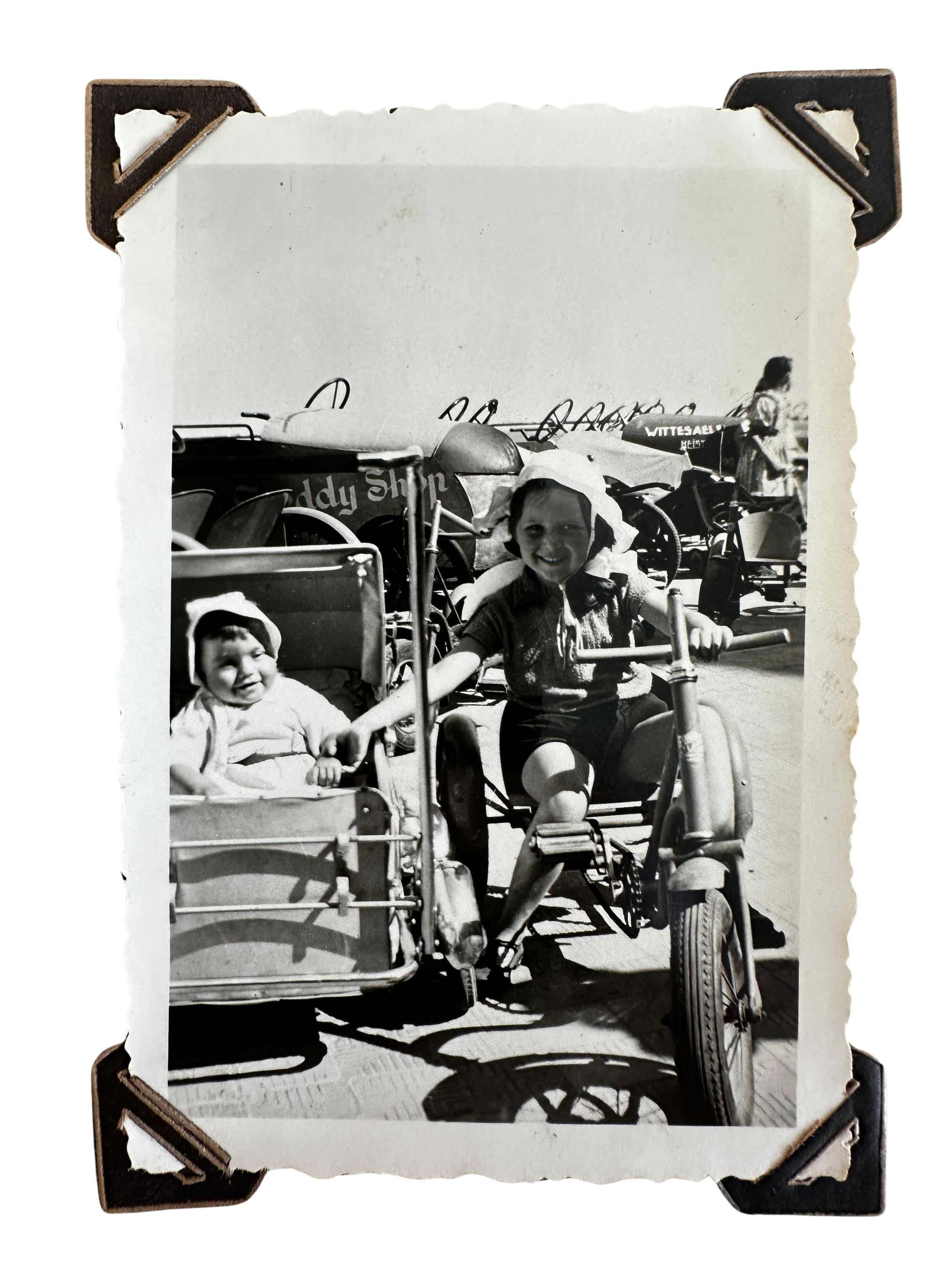 Twee meisjes op een fiets met een zijspan, op een strand met andere attracties en mensen op de achtergrond, zwart-wit foto.