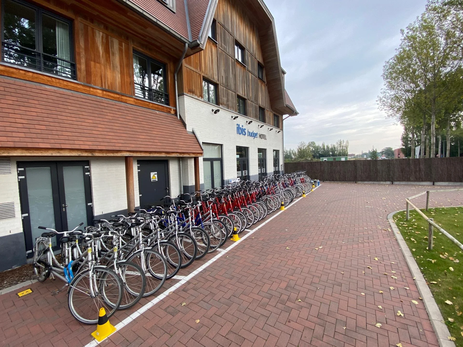Fietsparkeerplaats voor hotel met rijen fietsen, naast een gebouw met houten en witte gevels, en een hek met bomen op de achtergrond.
