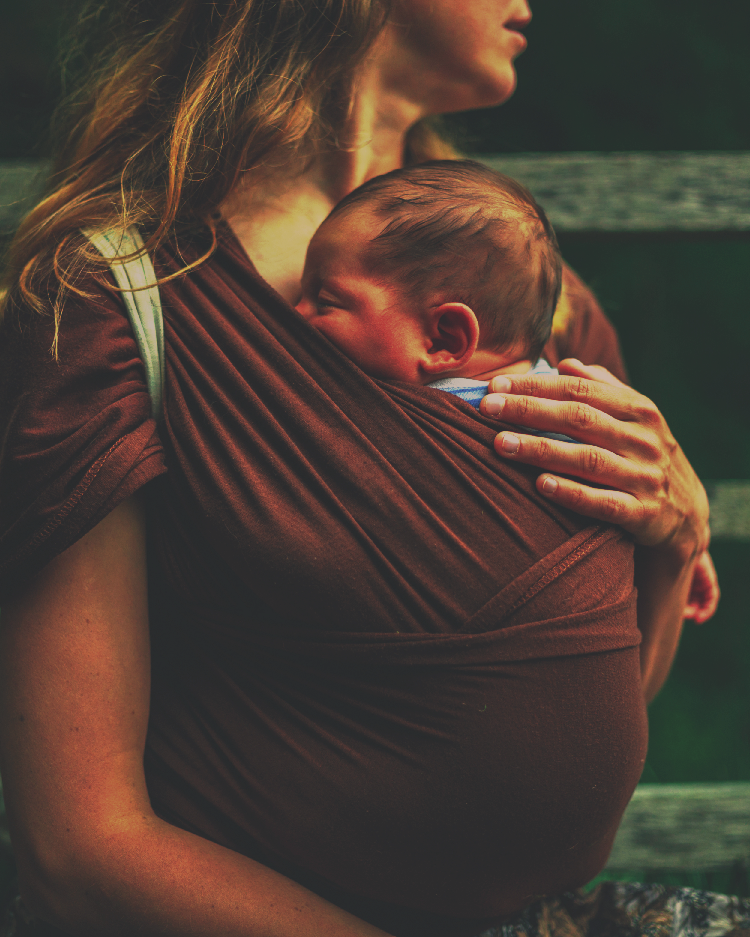 Mother carrying a sleeping baby in a brown wrap carrier outdoors.
