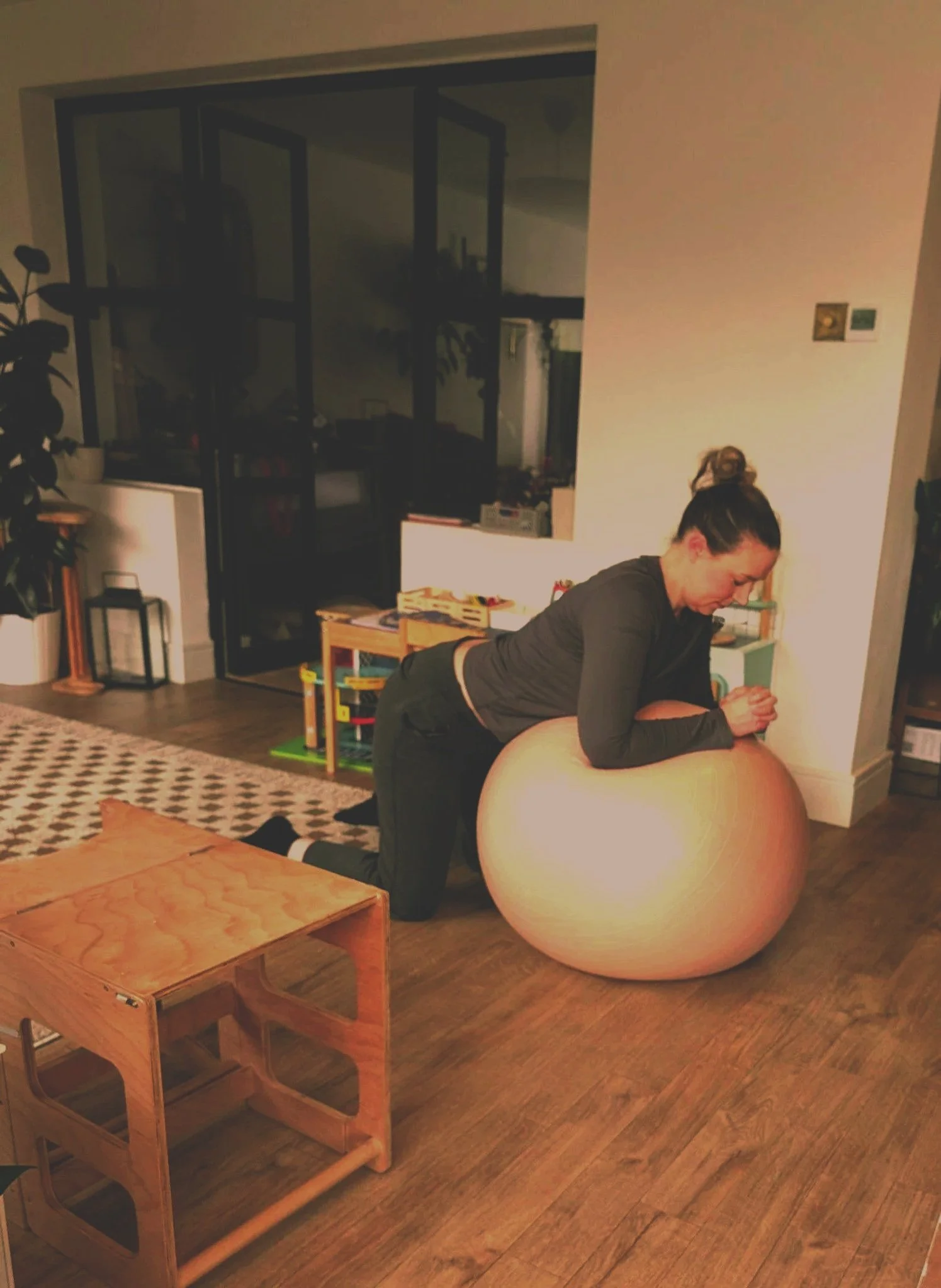 A woman in black workout clothes kneeling on a wooden floor, resting on her forearms on a large beige exercise ball inside a living room.