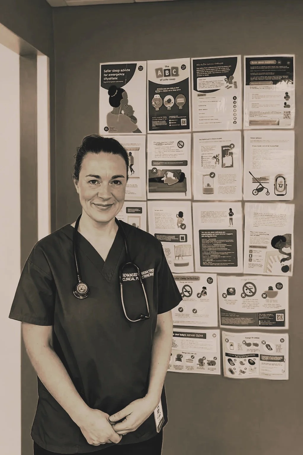 A female healthcare worker in scrubs and a stethoscope standing in front of a bulletin board with health information posters.