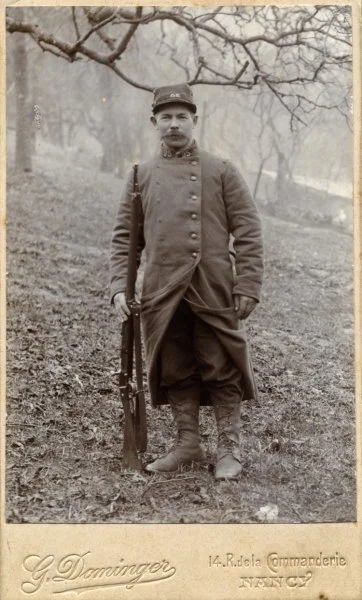 Un soldat debout dans un champ avec des arbres en arrière-plan, portant un uniforme militaire ancien, avec un fusil tenu à la main