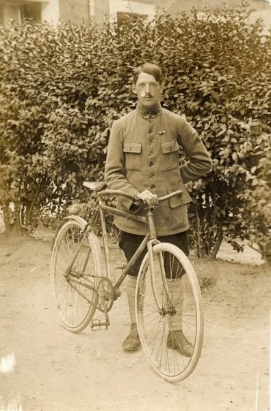 Homme en uniforme militaire posant avec un vieux vélo devant un buisson dense.