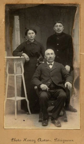 Une photographie en noir et blanc de trois personnes dans un studio, probablement une famille. Deux personnes sont debout à l'arrière, une femme à gauche et un homme à droite, tous deux en vêtements anciens. Un homme plus âgé est assis au centre, por