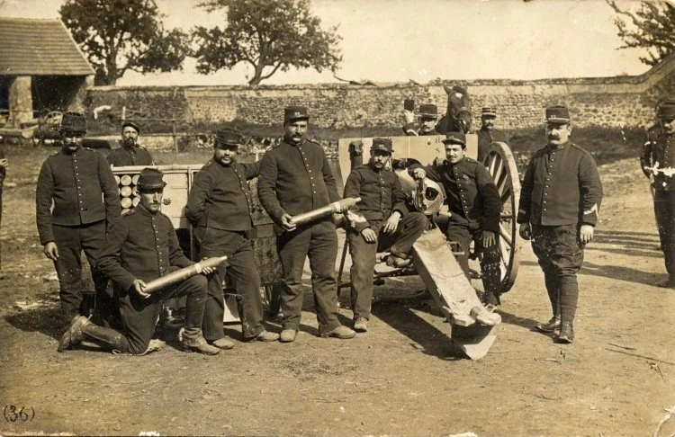 Groupe d'hommes en uniformes militaires posant avec un canon antique lors d'une journée en plein air, avec des arbres et un mur en arrière-plan.