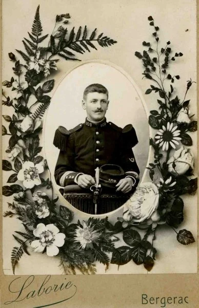 Portrait d'un homme en uniforme militaire, entouré de fleurs et de feuilles, dans un cadre oval.
