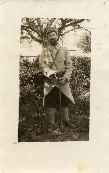 Homme en uniforme militaire avec un chapeau, debout dans un jardin avec des buissons et un arbre en arrière-plan, tenant un objet dans ses mains.