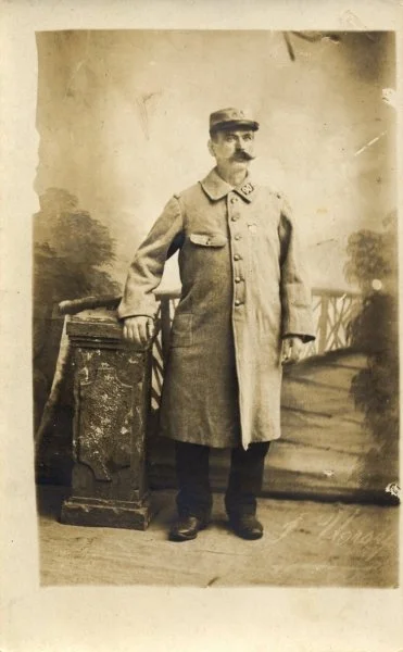Un homme habillé d'un long manteau, portant une casquette, posant à côté d'une petite table en bois dans un studio photo ancien.