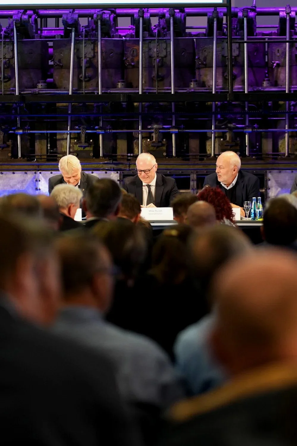 Drei Männer sitzen an einem langen Tisch bei einer Konferenz oder einem Meeting, mit einem Publikum im Vordergrund. Hinter ihnen befindet sich eine große technische Anlage oder Maschinerie mit purple Beleuchtung.