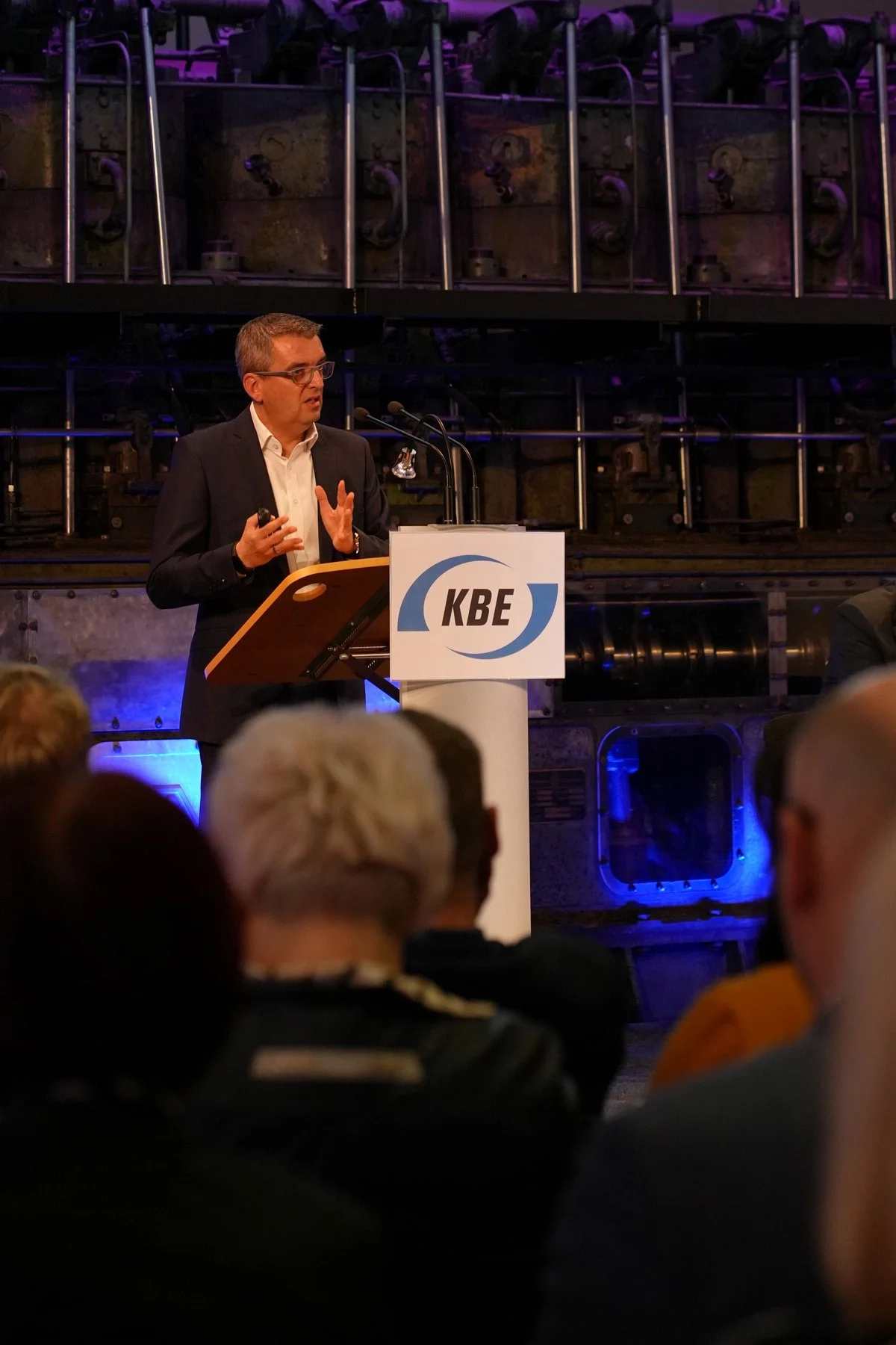 Unternehmensreportage - Redner am Podium mit gestikulierenden Händen vor Publikum, industrielles Ambiente - Businessfotografie Culet