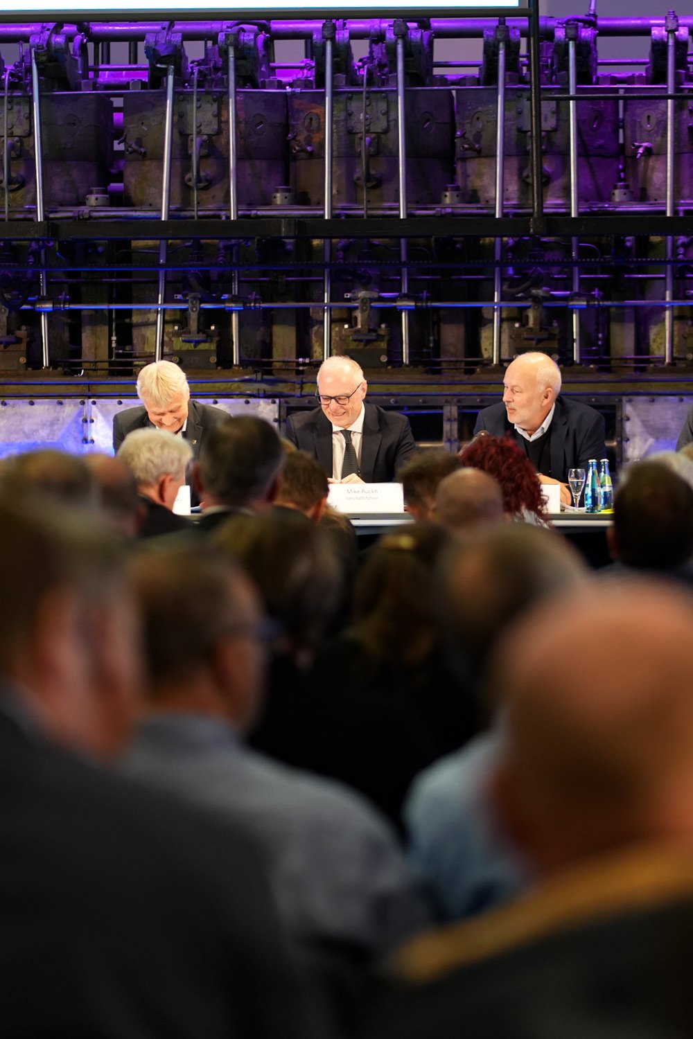 Unternehmensreportage - drei Redner lachen an einem Podiumstisch vor Publikum, industrielles Ambiente in lila Beleuchtung - Culet