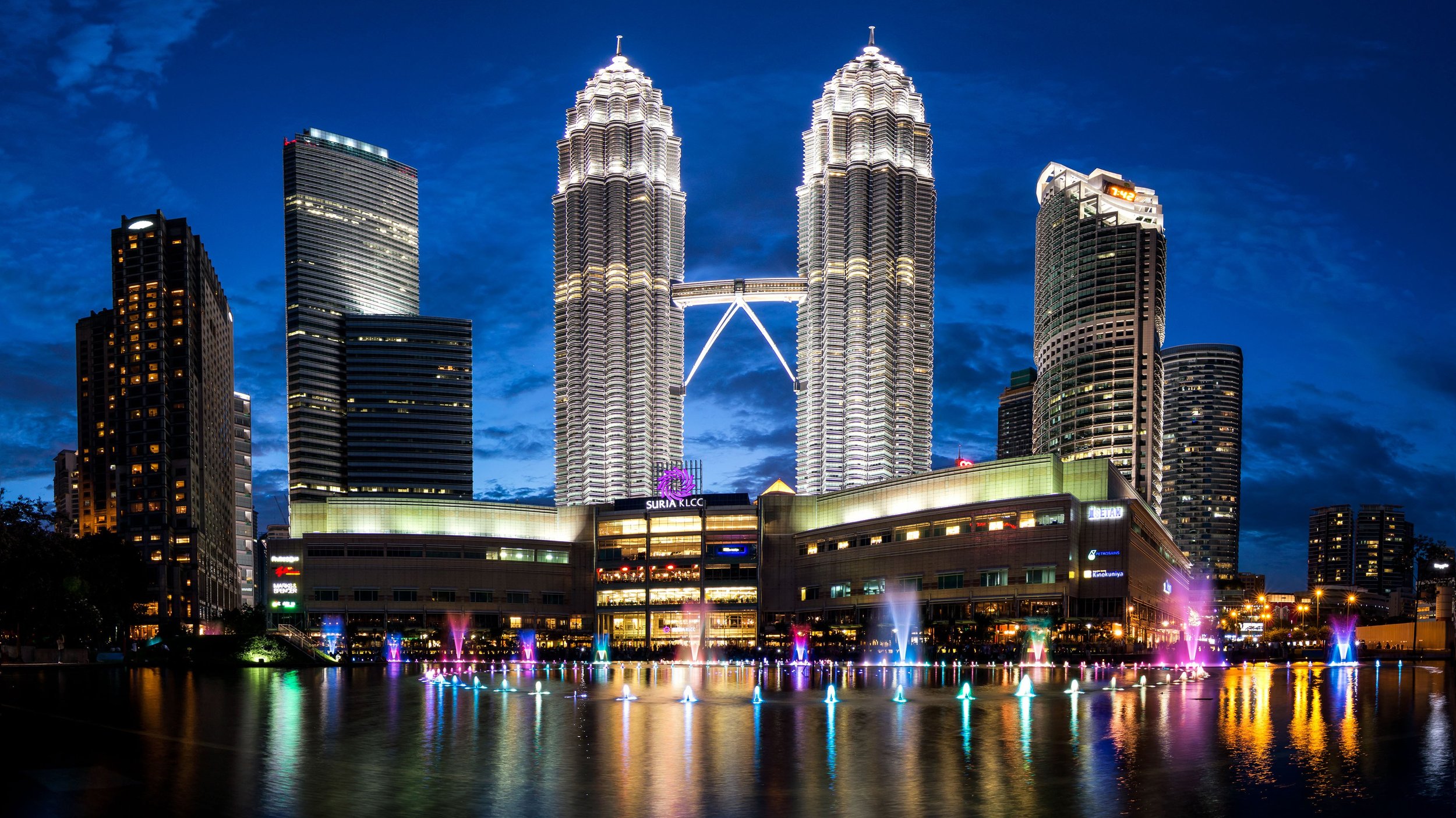 Night view of the Petronas Towers in Kuala Lumpur, Malaysia, illuminated with reflection in the water fountain below, surrounded by other tall city buildings.