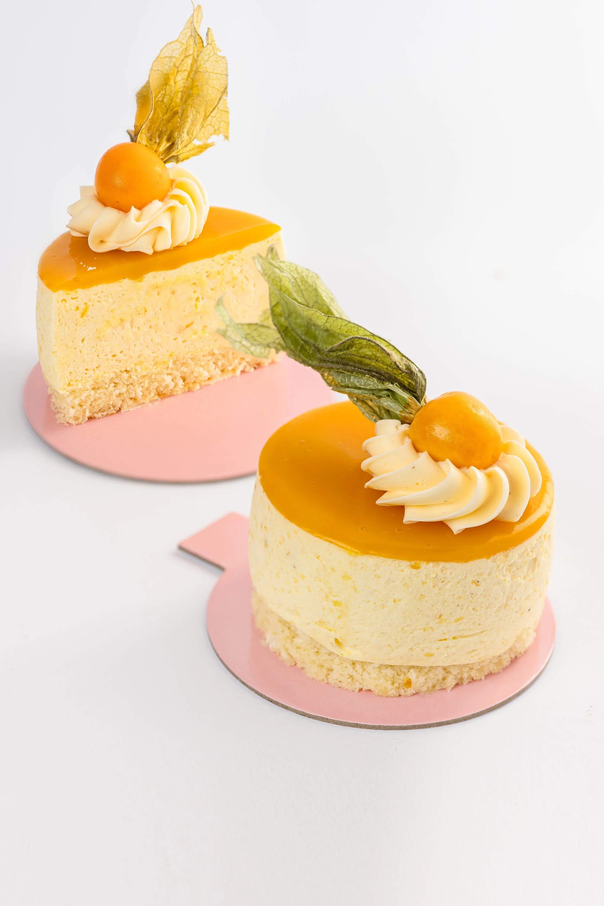 Zwei kleine, runde Käsekuchen mit Mango-Glasur, garnierte mit Mango-Physalis und Sahne auf einem pinken Untergrund.