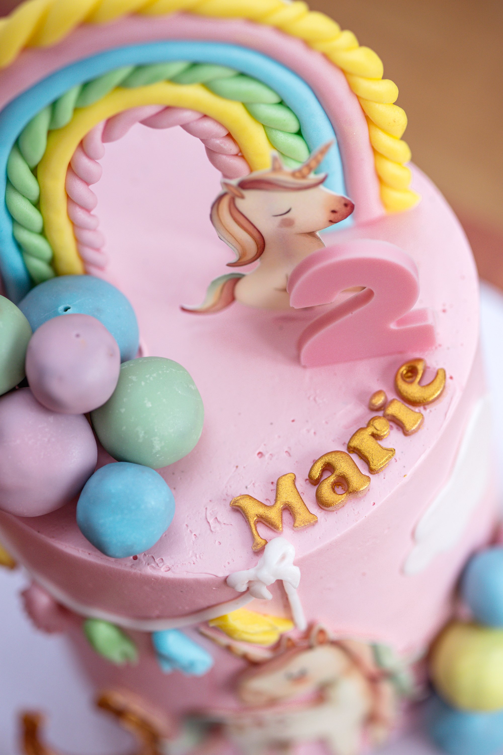 Ein rosa Geburtstagstorte mit Einhorn- und Regenboga-Details, bunten Kugeln, einer großen pinken Zahl 2, und dem Namen Mara in Goldbuchstaben.