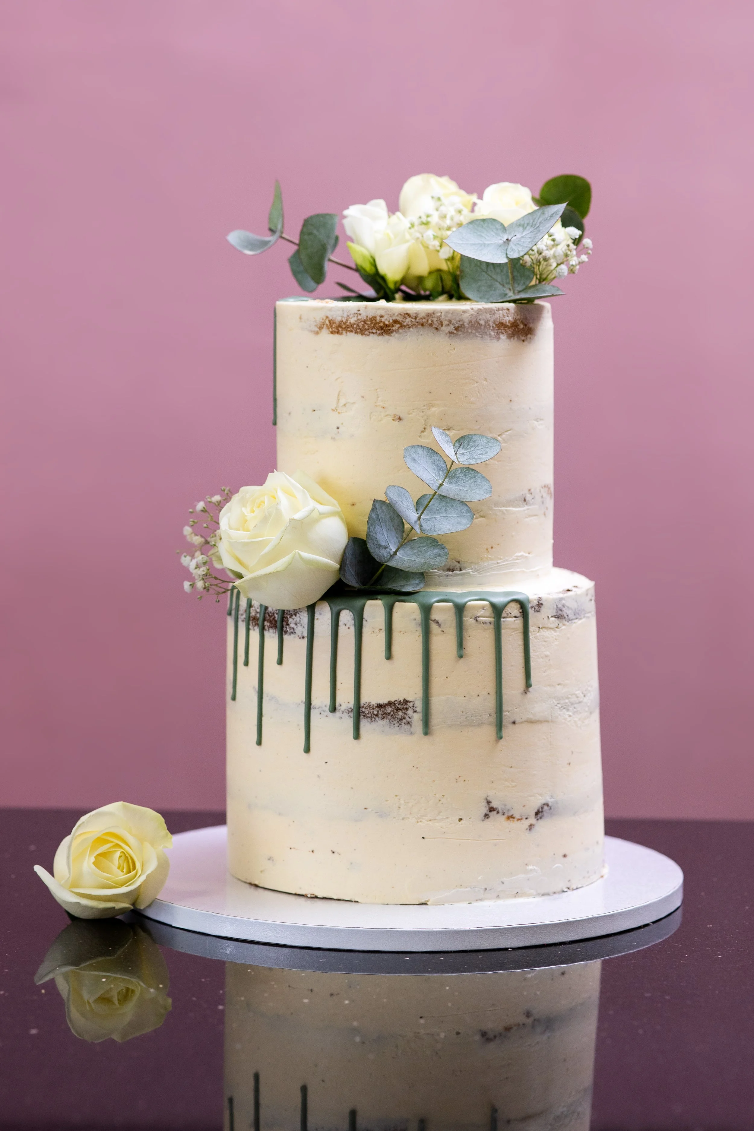 Ein zweistöckiger wedding cake mit weißer Zuckerglasur, grüner Kakaoguss, weißen Rosen und grünen Eukalyptusblättern auf einem schwarzen Tisch vor rosa Hintergrund.