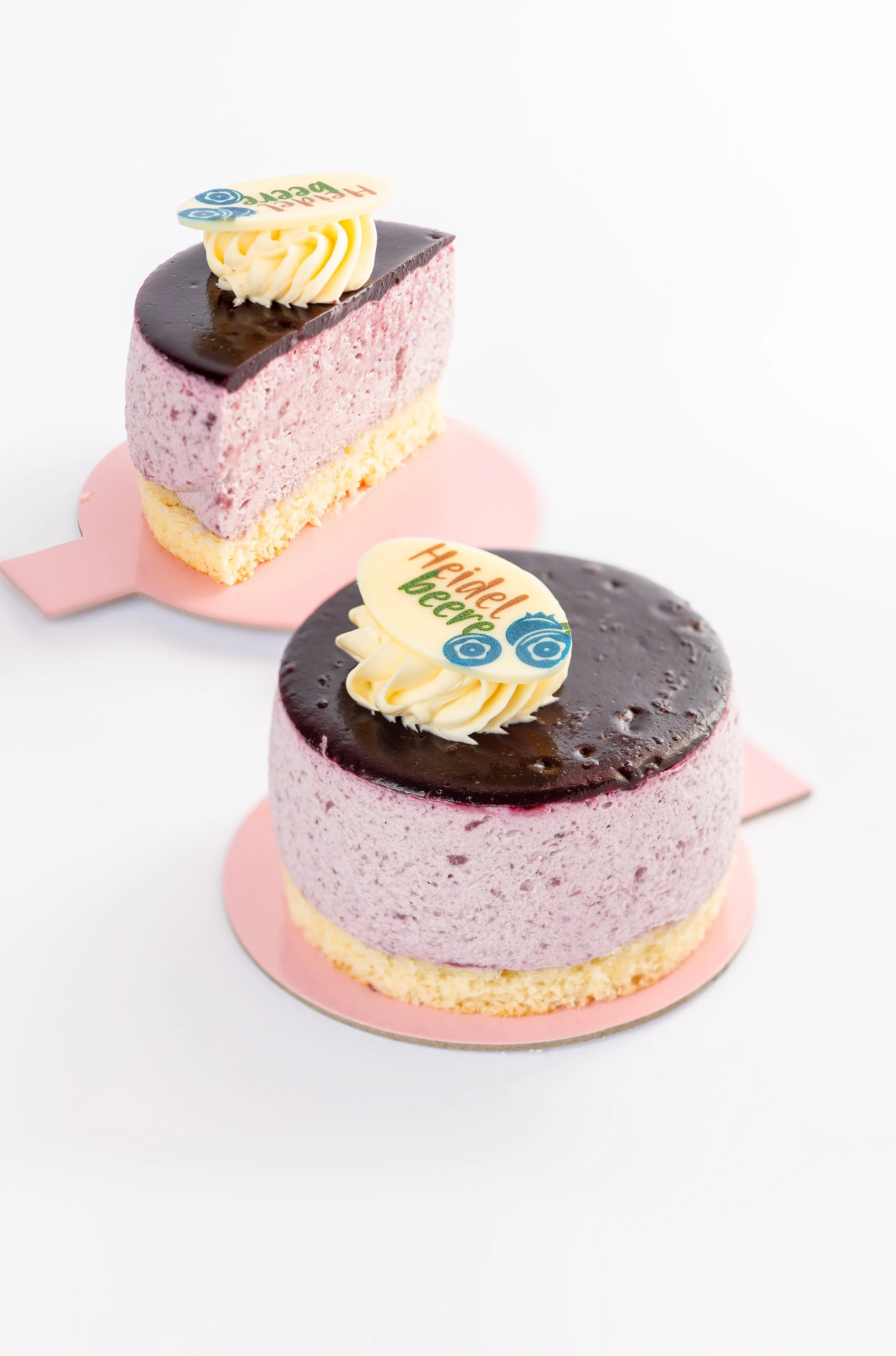 Zwei kleine, runde, pinke Kuchen mit dunkler Schokoladenglasur und Dekorationen auf weißen Unterlagen.