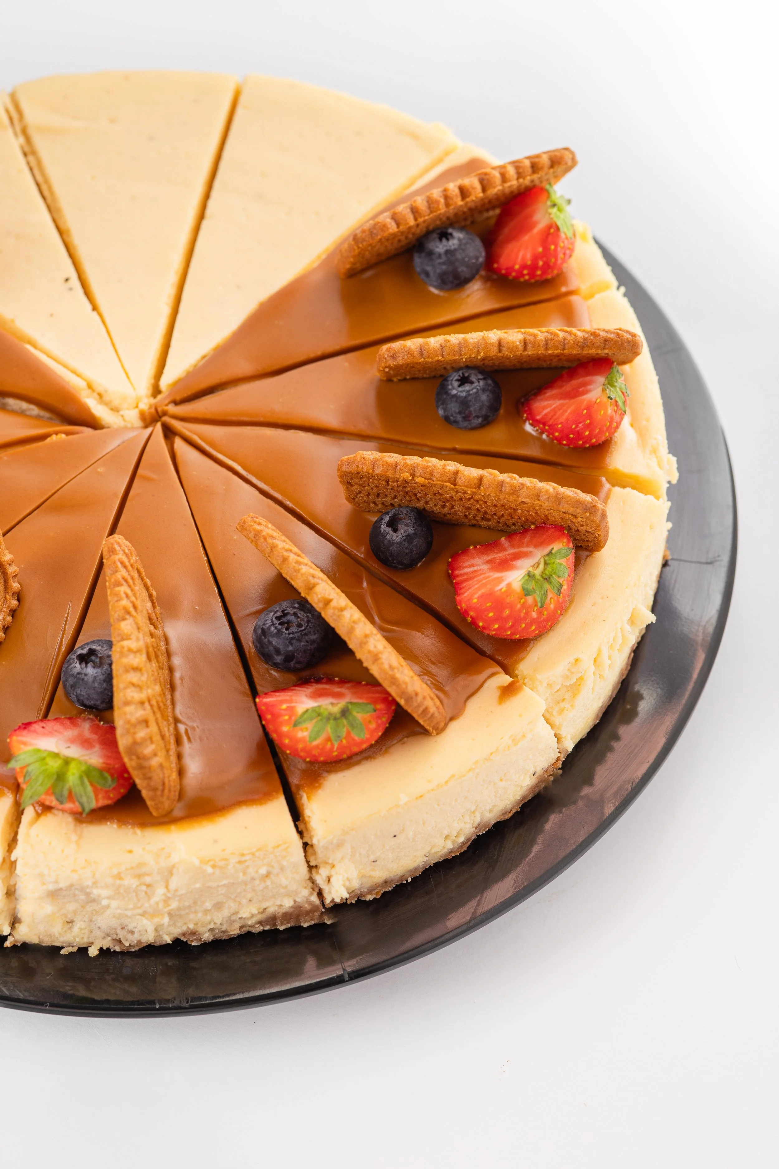 Käsekuchen mit caramelisierten und rund verzierten Keksstücken, Erdbeeren, Heidelbeeren und weiteren Keksen auf einem schwarzen Teller