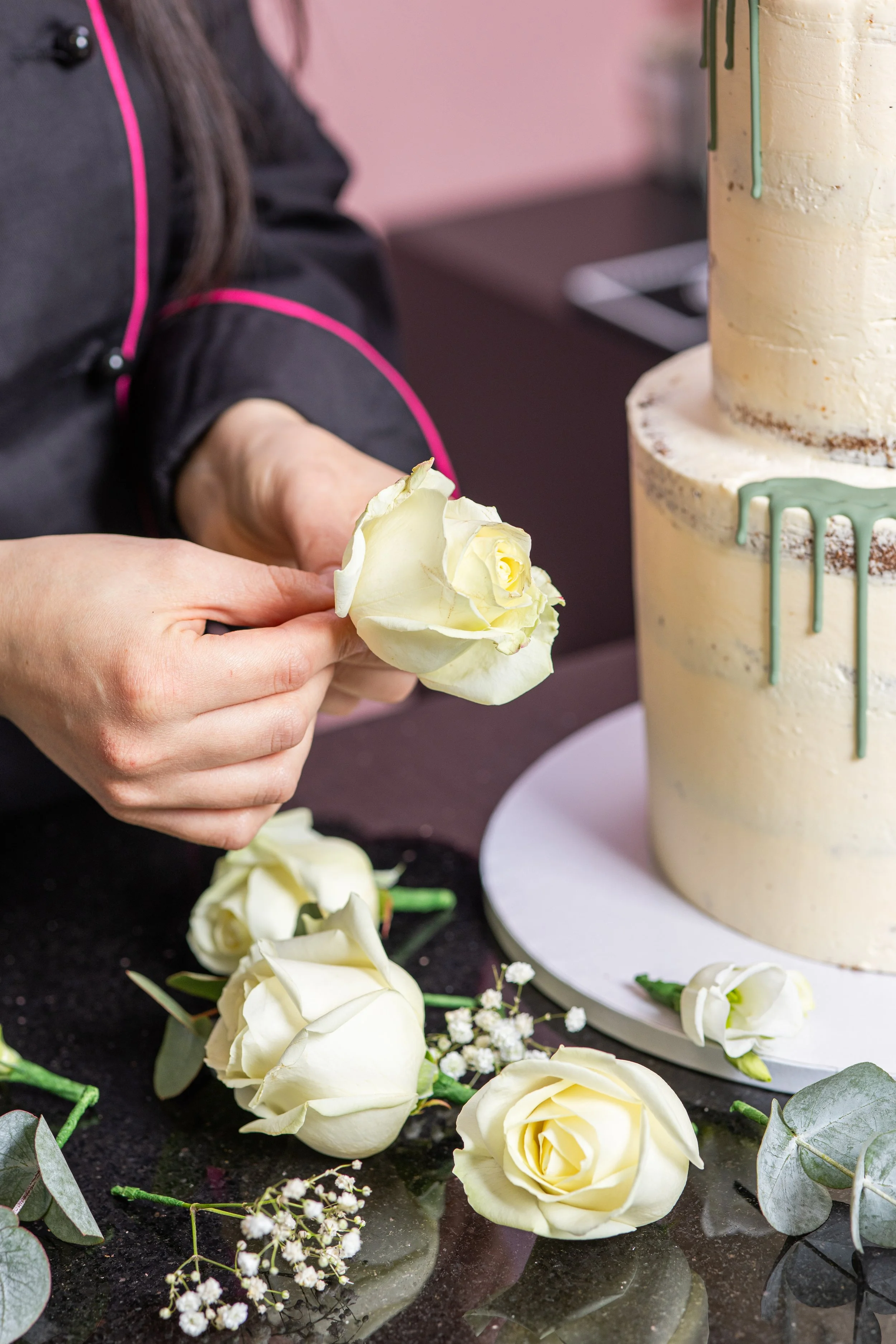 Person dekoriert eine zweistöckige weiße Hochzeitstorte mit weißen Rosen und Eukalyptusblättern.