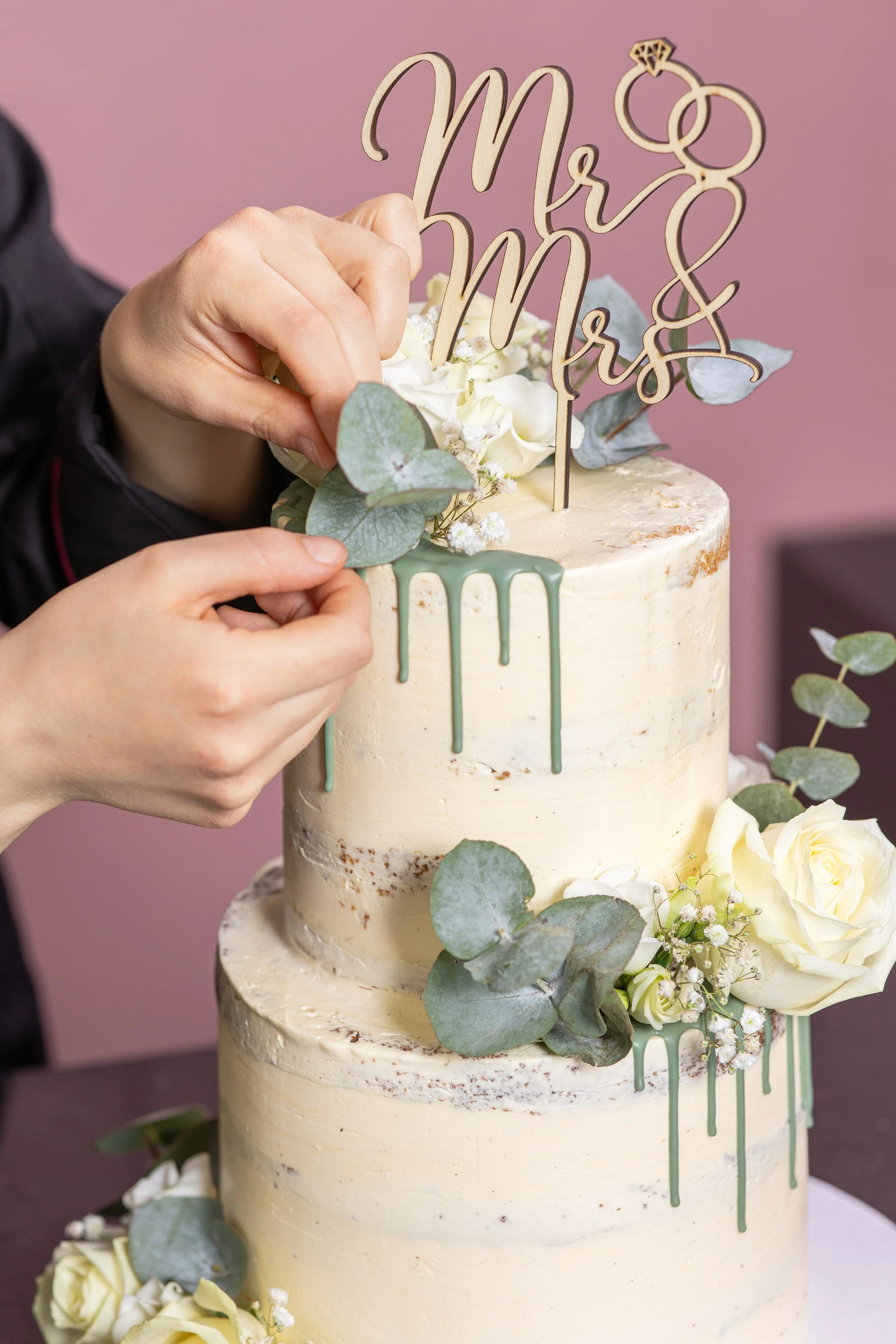 Hand befestigt eine Dekoration mit den Worten "Mr & Mrs" auf einer Hochzeitstorte, die mit weißen Blumen und grünen Blättern verziert ist.