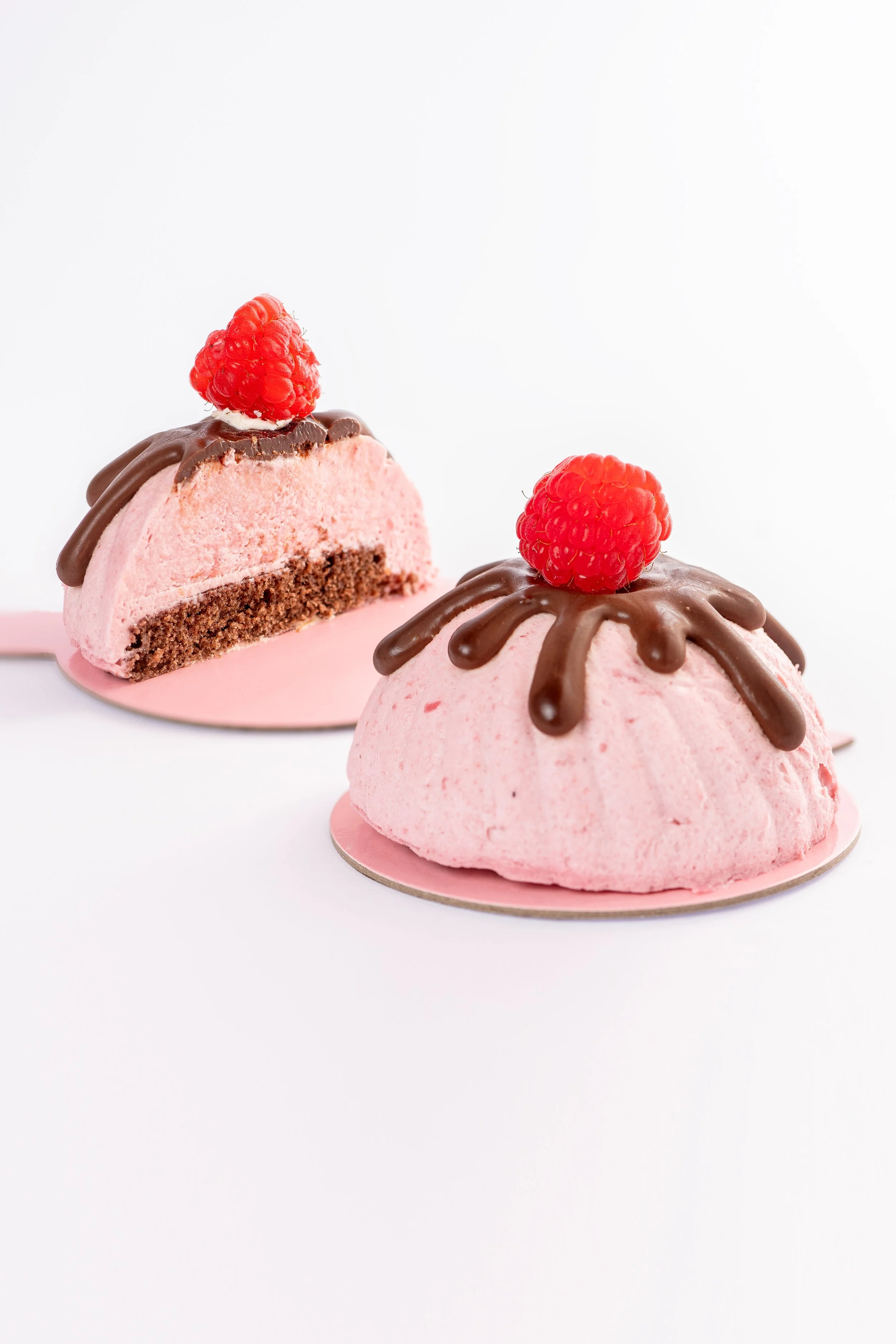 Zwei pinke, runde Dessertringe mit Schokoladenglasur und Himbeern auf einem weißen Hintergrund.