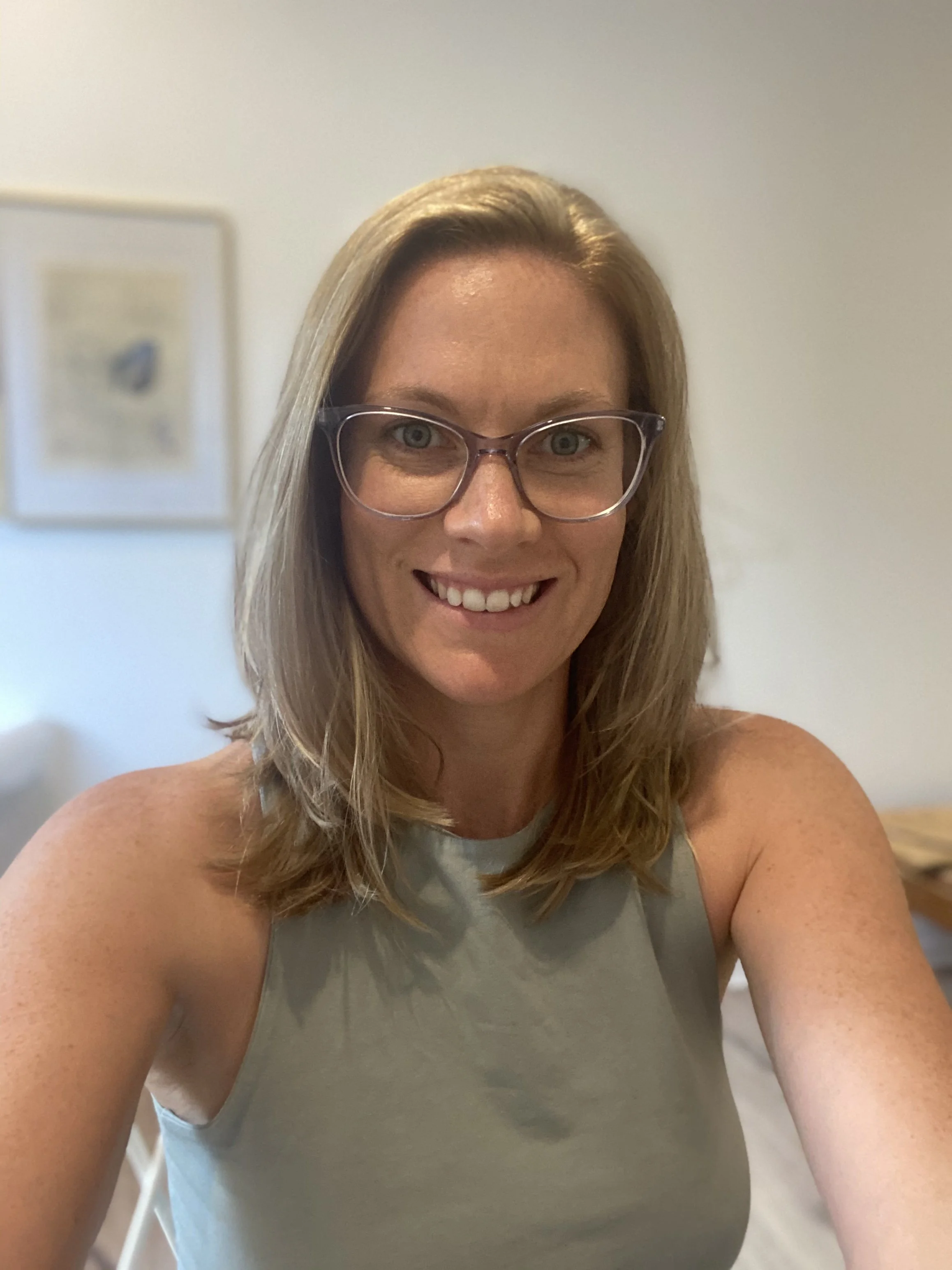 A woman with shoulder-length light brown hair, wearing glasses and a sleeveless gray top, smiling at the camera in a well-lit room