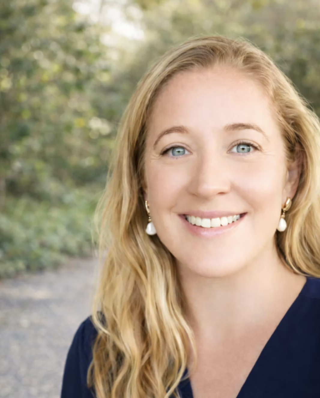 Close-up of a smiling woman with long, wavy blonde hair, wearing pearl earrings and a navy top, outdoors with blurred green foliage in the background.