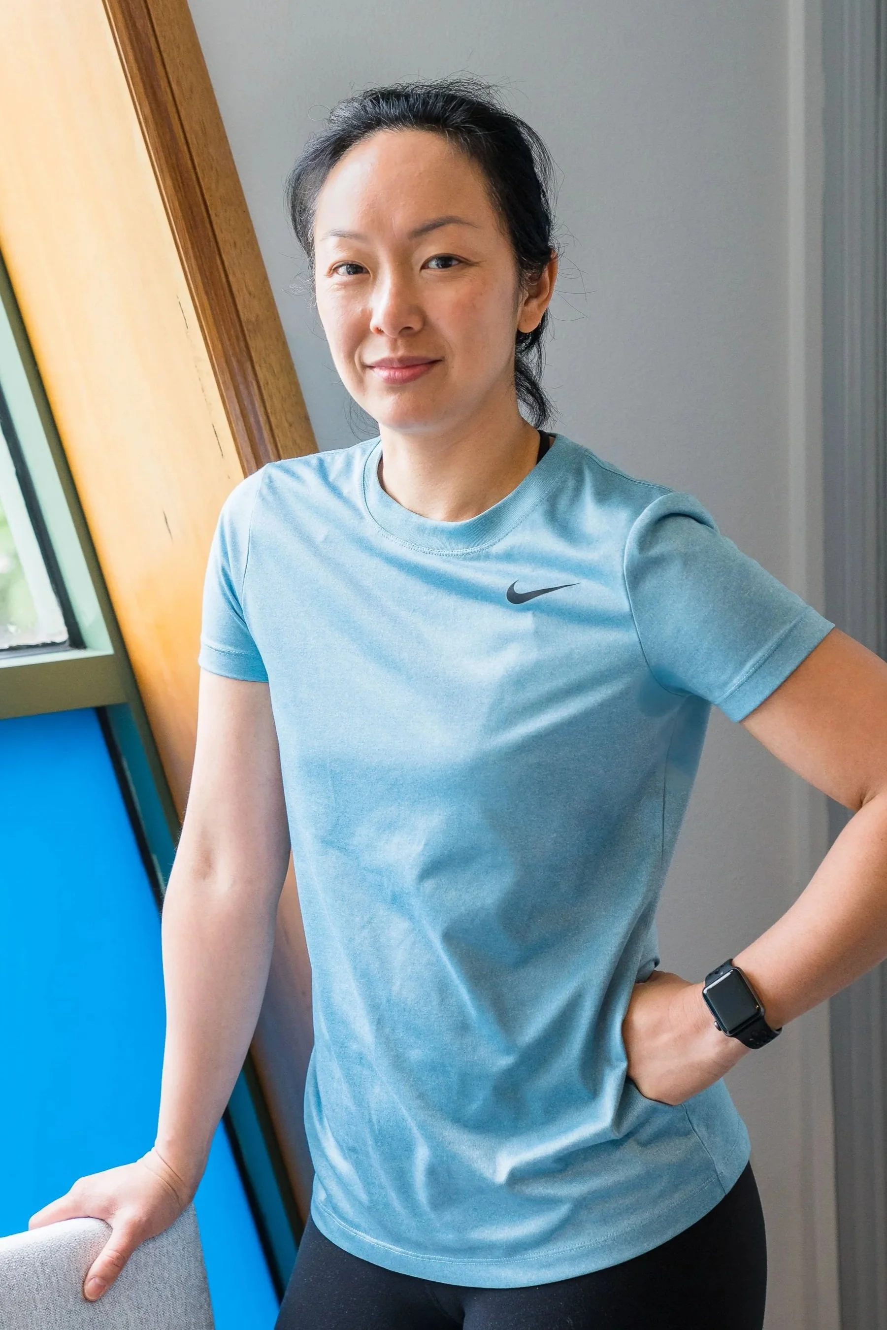 A massage therapist wearing a light blue Nike athletic shirt and a smartwatch, standing indoors near a blue wall and a wooden window frame.
