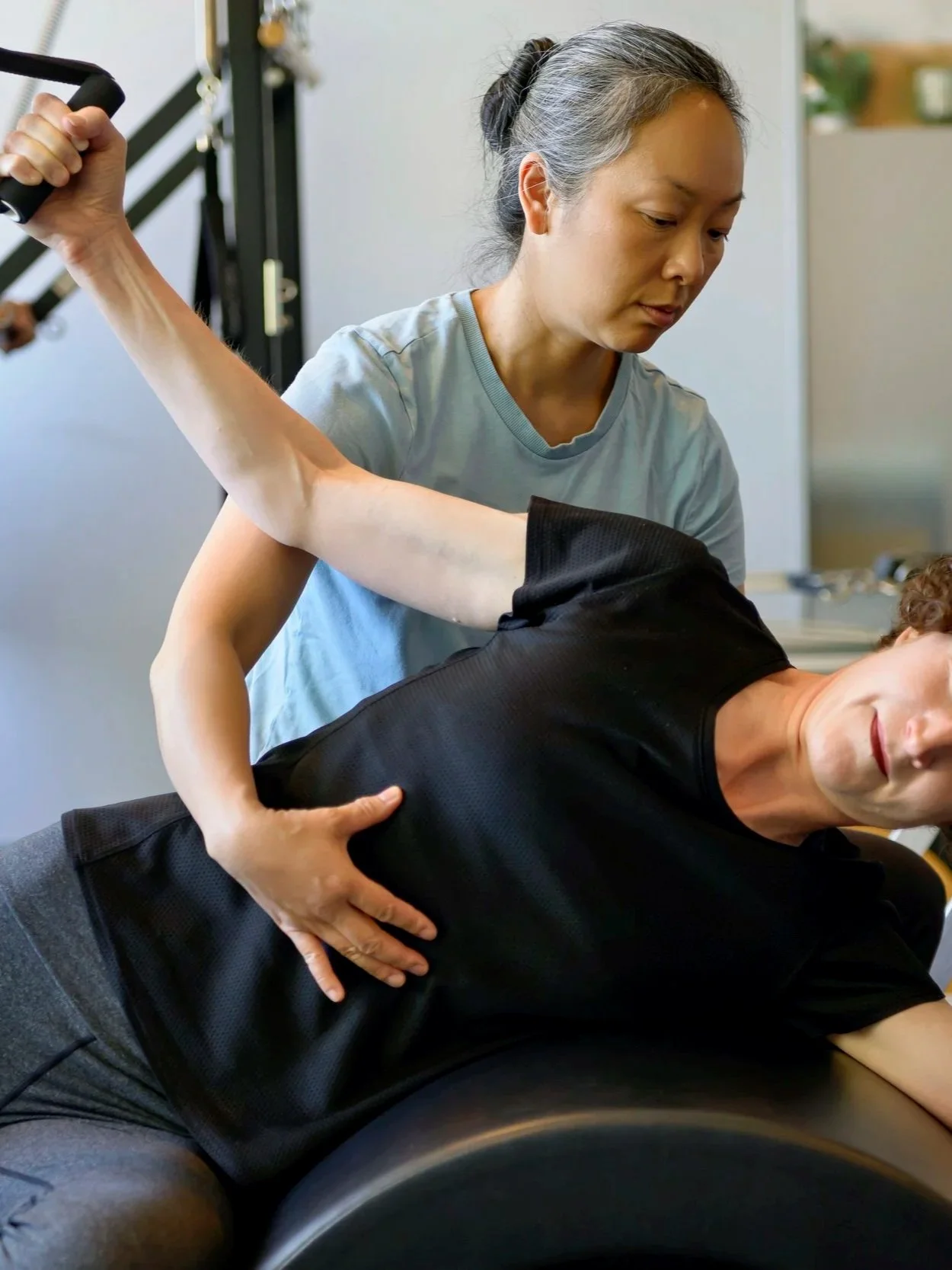 A clinical Pilates instructor guiding a client through exercises over an arc barrel, with another person visible in the background.