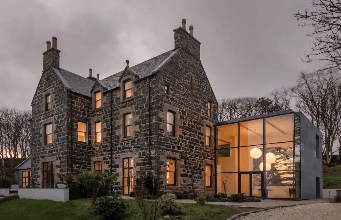 A stone castle-style house with multiple chimneys and a modern glass extension. It has several lit windows and is surrounded by landscaping with trees and grass during dusk or evening.
