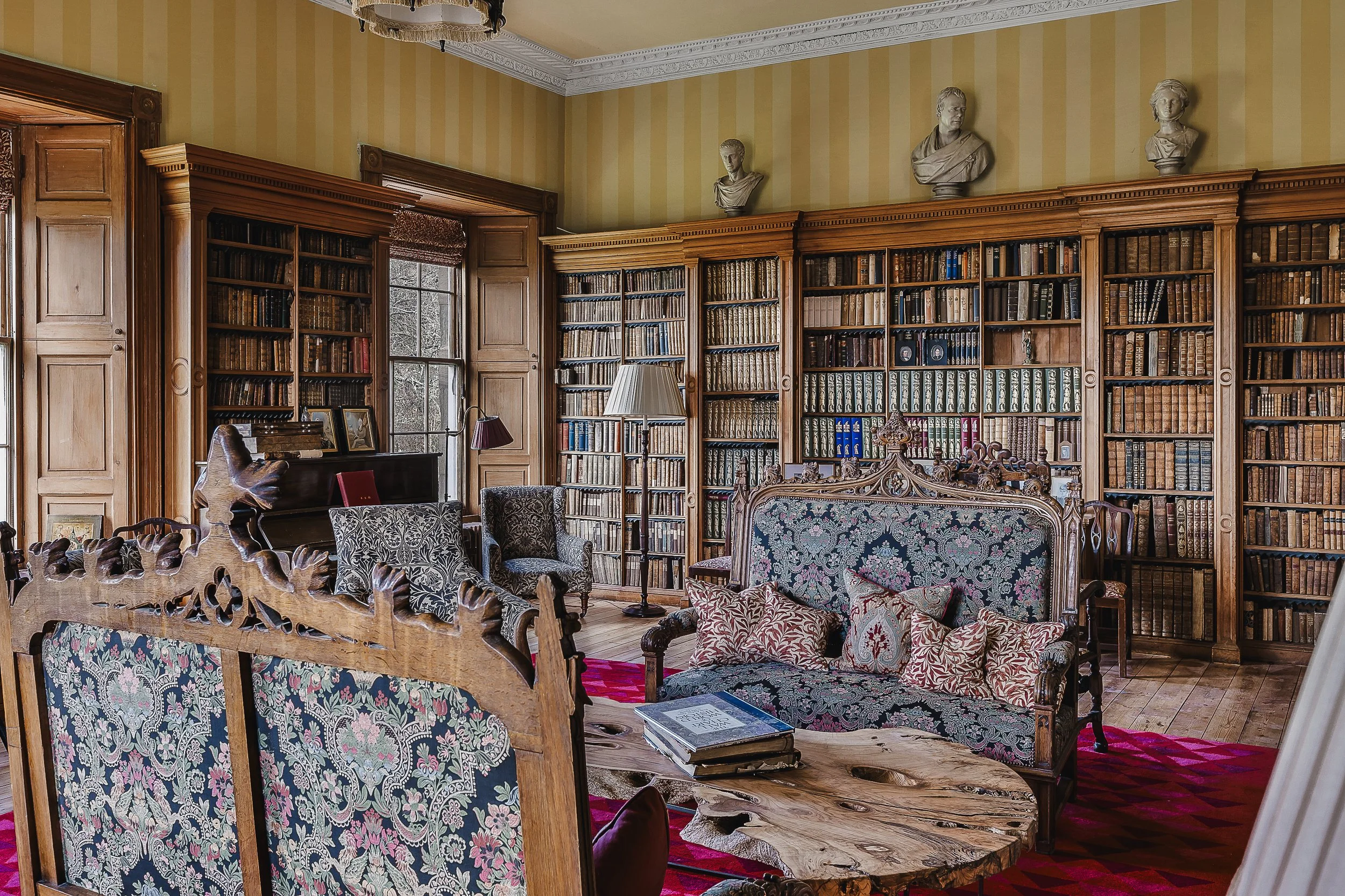 Elegant library room with tall wooden bookshelves filled with old books, bust sculptures on top, large windows with wooden shutters, antique chairs with floral upholstery, a carved wooden table, and a red rug.