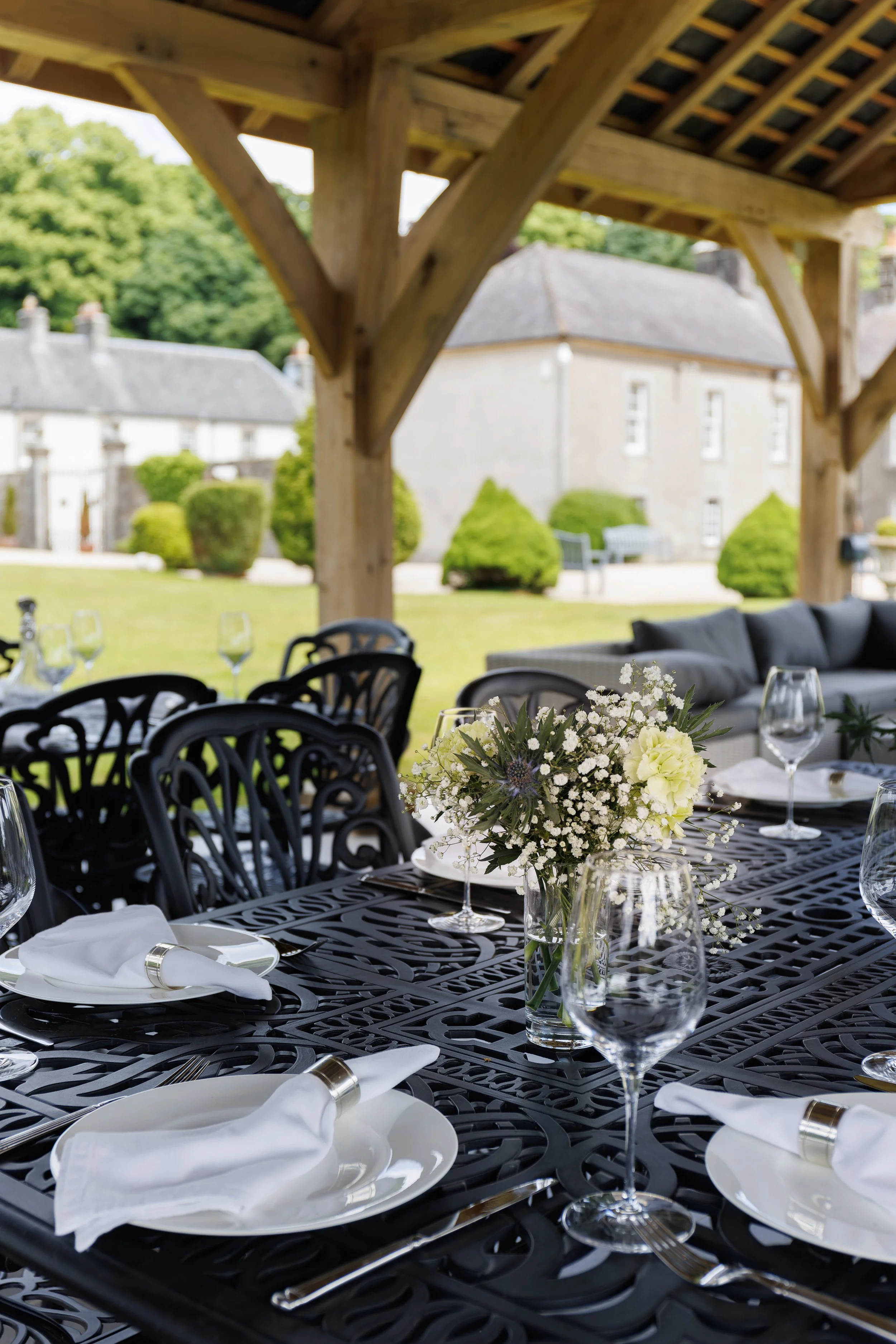 An outdoor dining setup featuring a black metal table with white plates, silver cutlery, white napkins, and glassware. A floral centerpiece with white flowers sits at the center of the table. In the background, there is a stone building, green shrubs