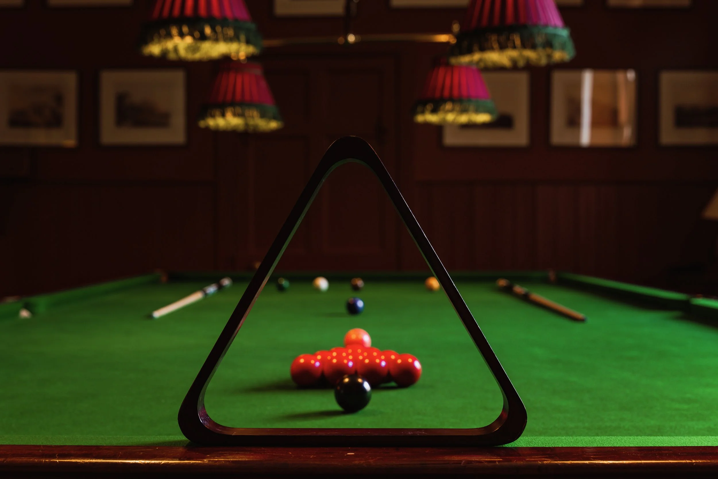 A close-up view of a billiards or pool table with a cue ball, colored balls, and a triangle rack in the foreground, set in a dimly lit room with framed pictures on the dark wooden wall and hanging lamps with red and green shades overhead.