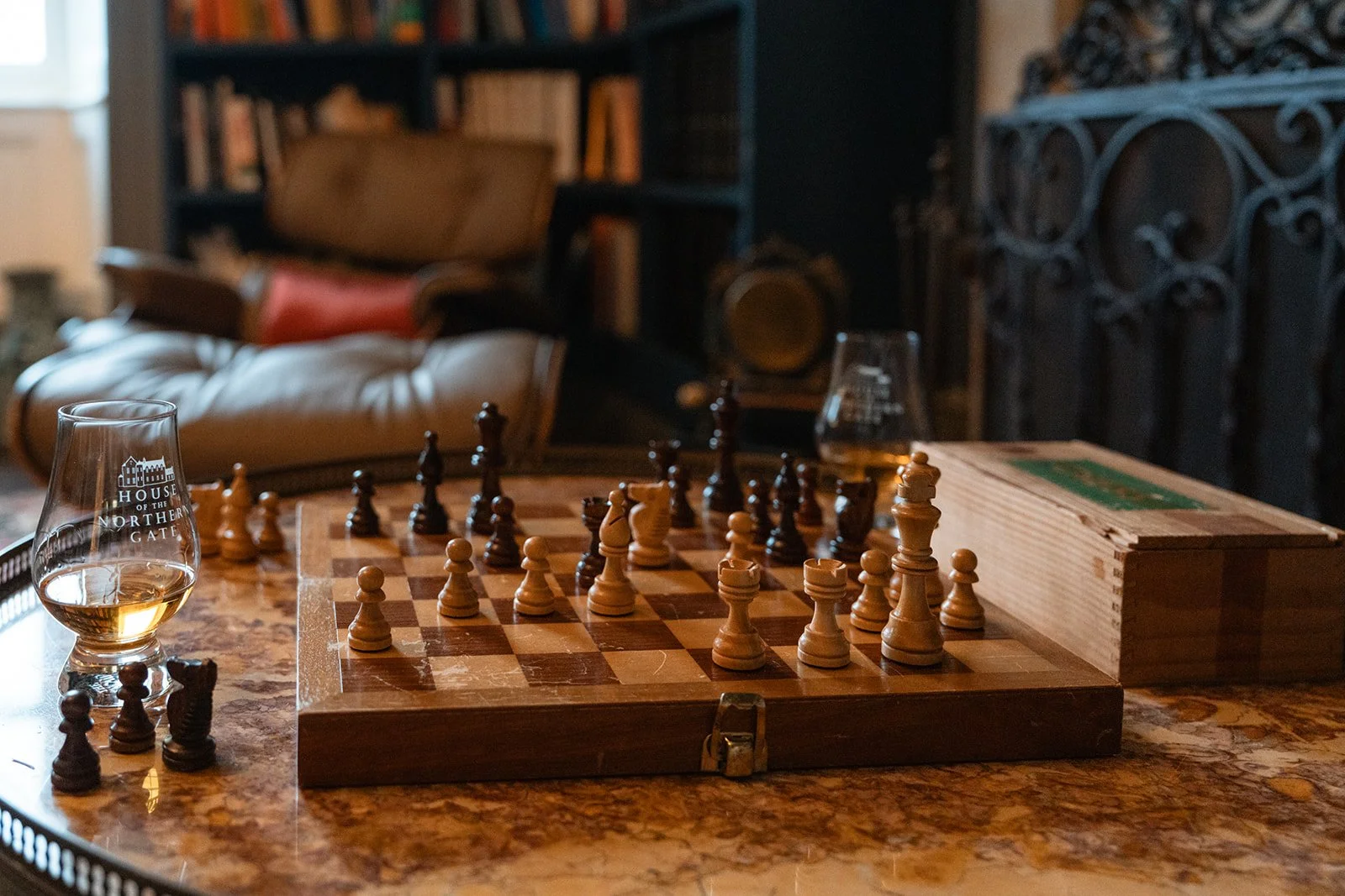A wooden chessboard with chess pieces set up for a game, a glass of whiskey, and a wooden box on a marble table in a cozy living room with a leather armchair, a bookshelf, and a fireplace in the background.