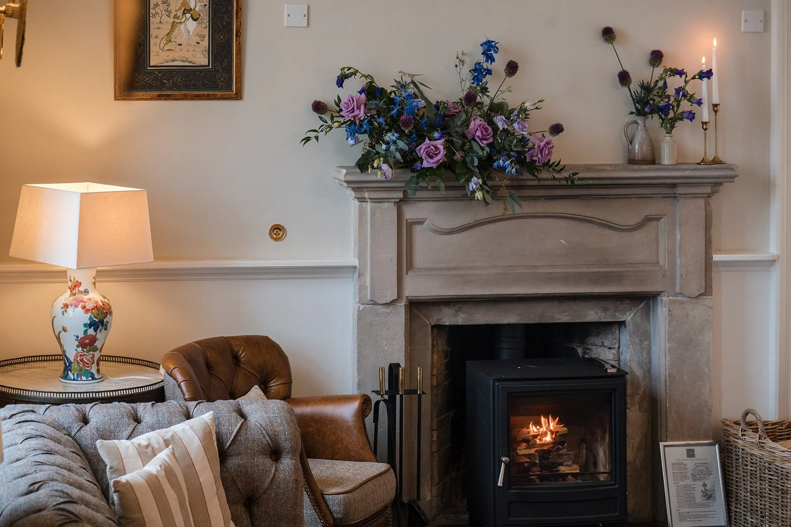 Living room with a stone fireplace, decorated with a large floral arrangement on the mantel. To the left, there is a side table with a floral lamp, a leather armchair, and a tufted sofa with striped pillows. On the mantel, there are candles and vases