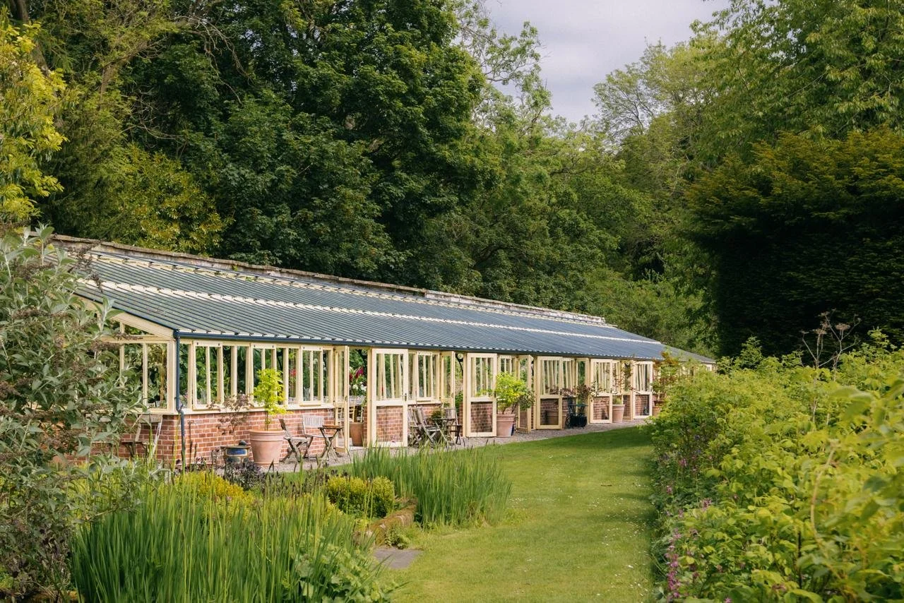 A greenhouse with a metal roof and glass walls situated in a lush garden with green plants and trees.