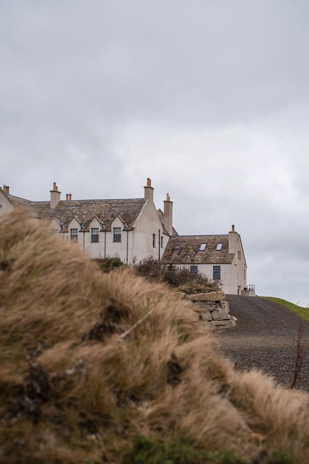 Historic white buildings with sloped roofs on a hilltop under a cloudy sky, foreground features tall grass and rocks.